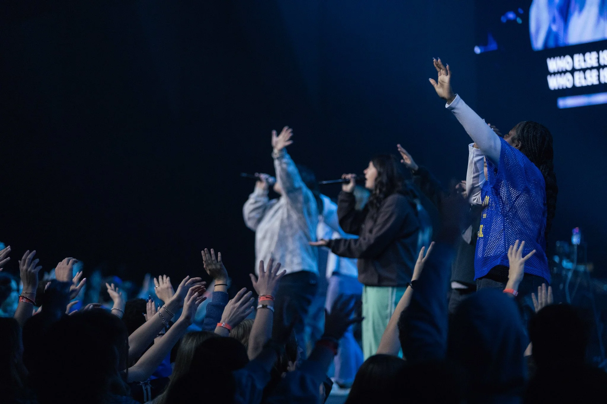 A group of performers on stage singing into microphones, with an audience reaching hands up towards them.