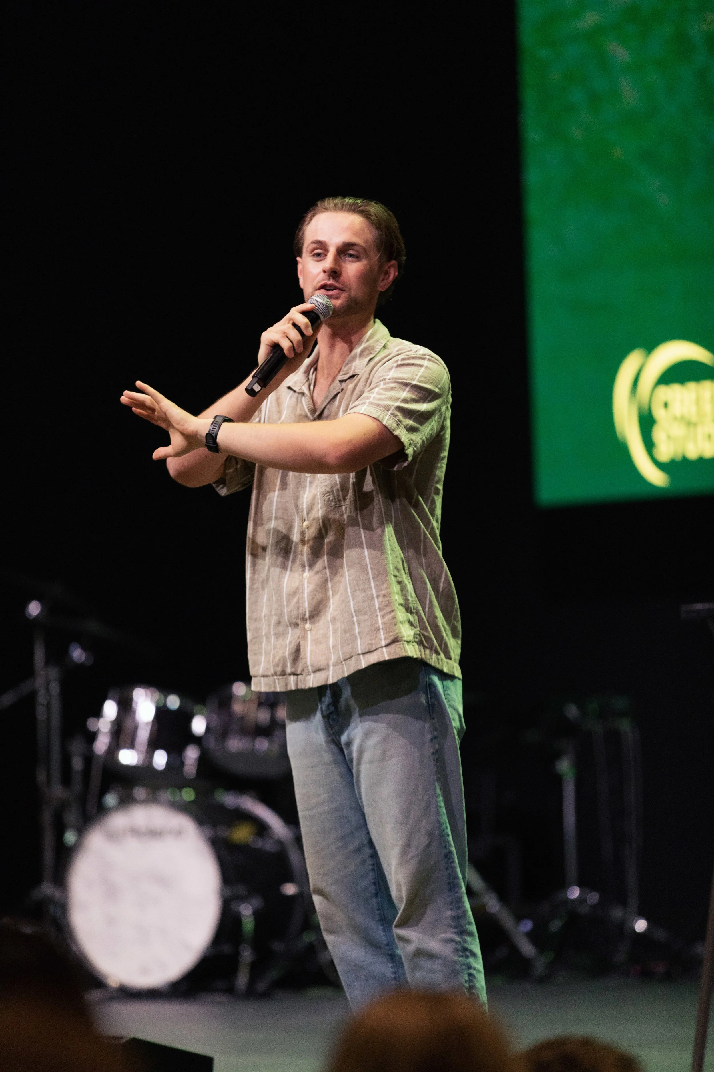 A young man with light brown hair, wearing a beige striped shirt and jeans, speaking into a microphone while gesturing with his left hand on stage with musical instruments in the background and a green presentation screen to his right.