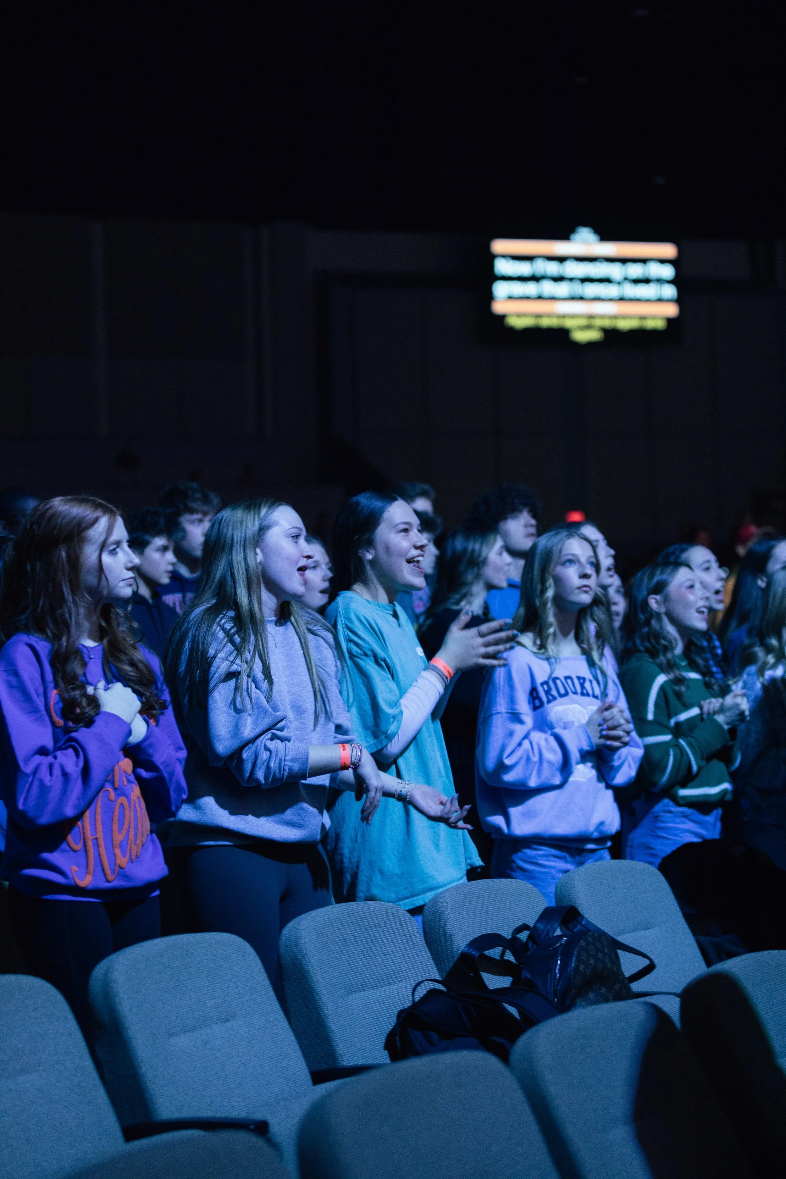 Group of young people standing and smiling in a dimly lit auditorium or conference hall, with empty chairs in the foreground and a screen with blurry text in the background.