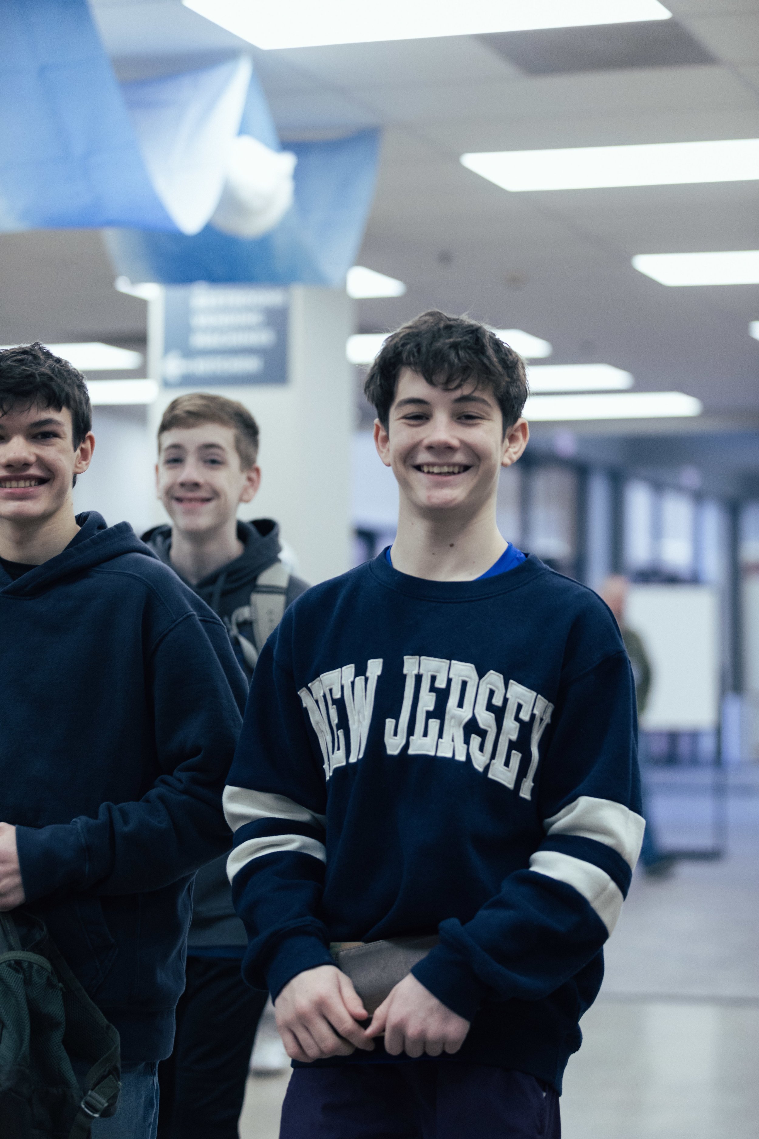 Three teenage boys standing together in a brightly lit indoor space, smiling at the camera. One boy in the front wears a navy sweatshirt with 'New Jersey' printed on it, while the other two boys are partially visible on either side. The background shows a hallway and ceiling lights.