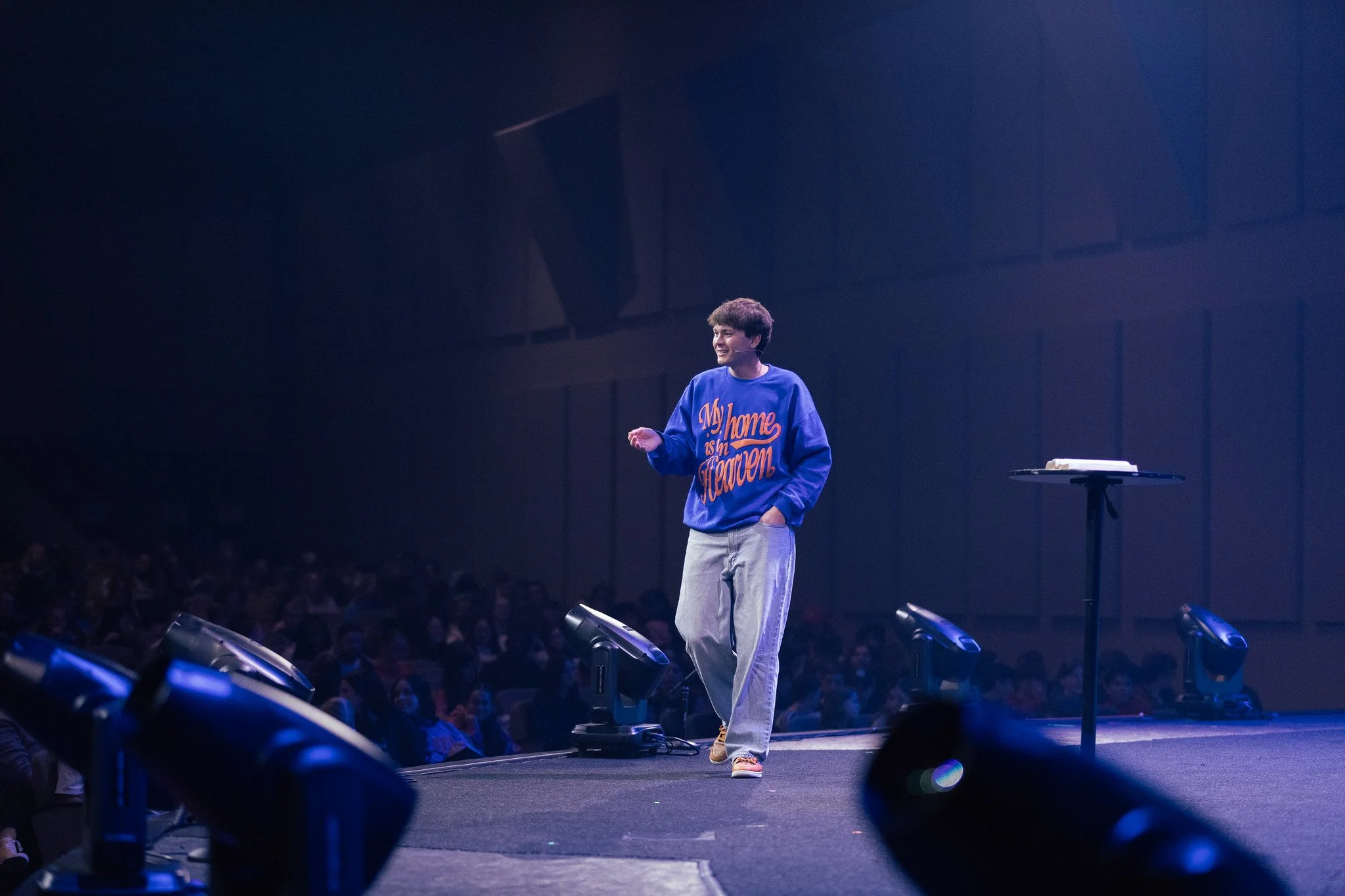 A young man standing on a stage, smiling, wearing a blue sweatshirt with orange text that says "My home is in Heaven," and light-colored pants, with a microphone headset, during a presentation or speaking event. There is a small table nearby with some objects, and the stage is lit with purple and blue lighting.