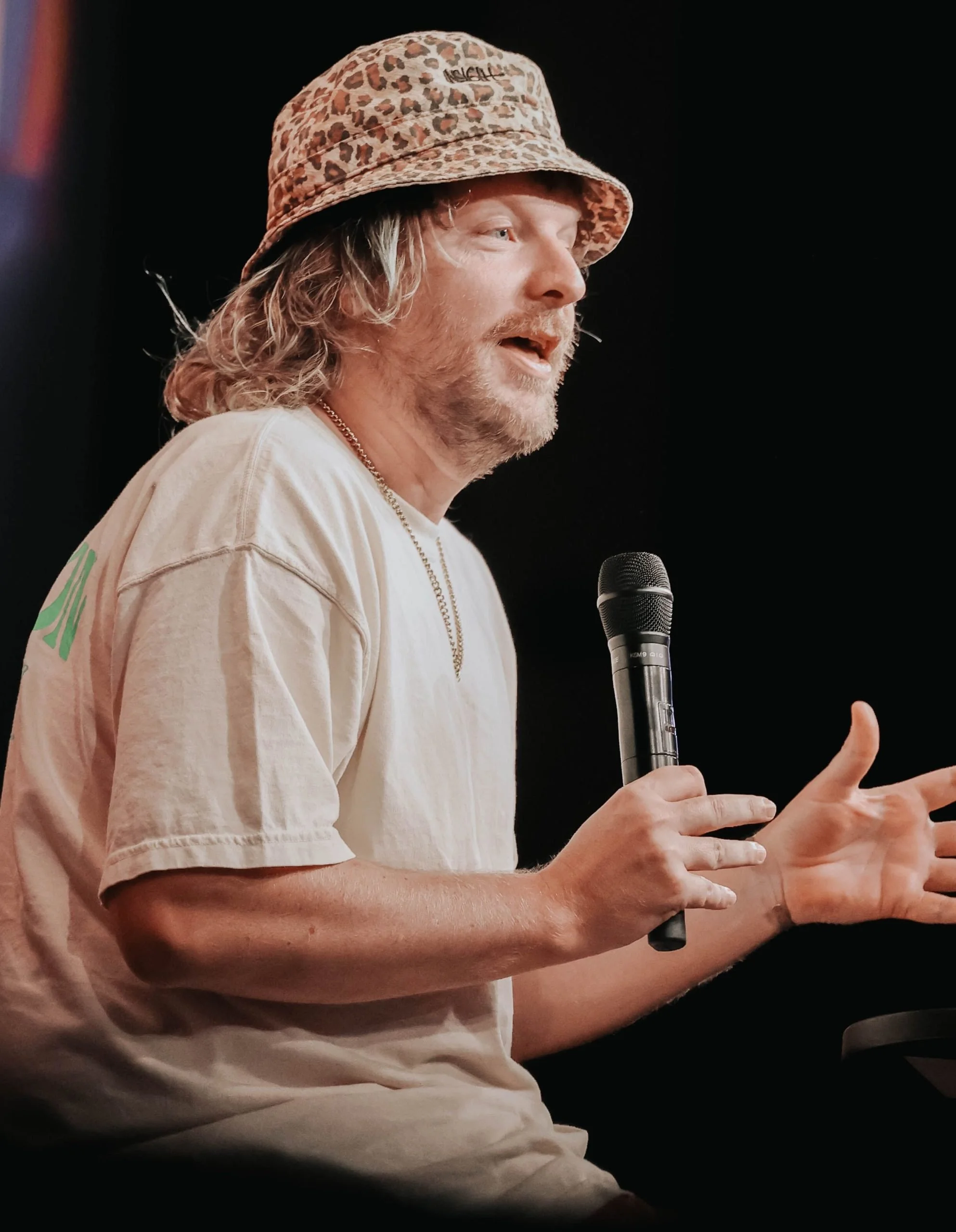 A man with long blond hair and a beard speaking into a microphone while gesturing with his hands, wearing a leopard print bucket hat and a beige t-shirt.