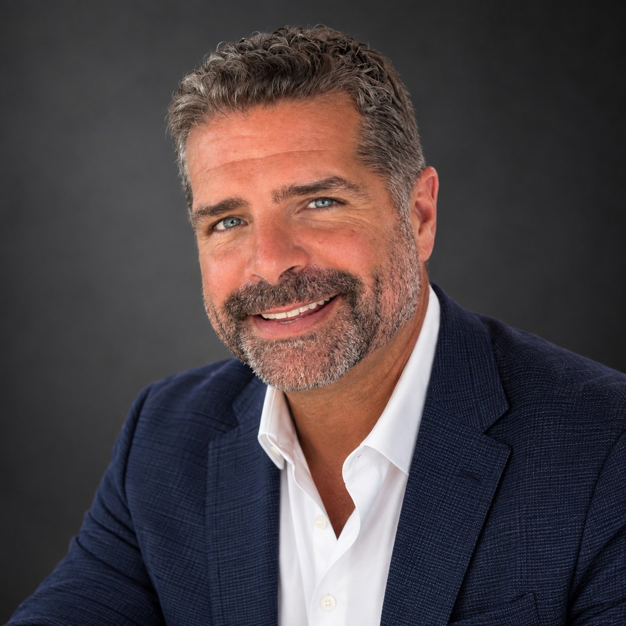 Headshot of a middle-aged man with salt-and-pepper hair and beard, smiling, wearing a navy blazer and white shirt against a dark grey background.