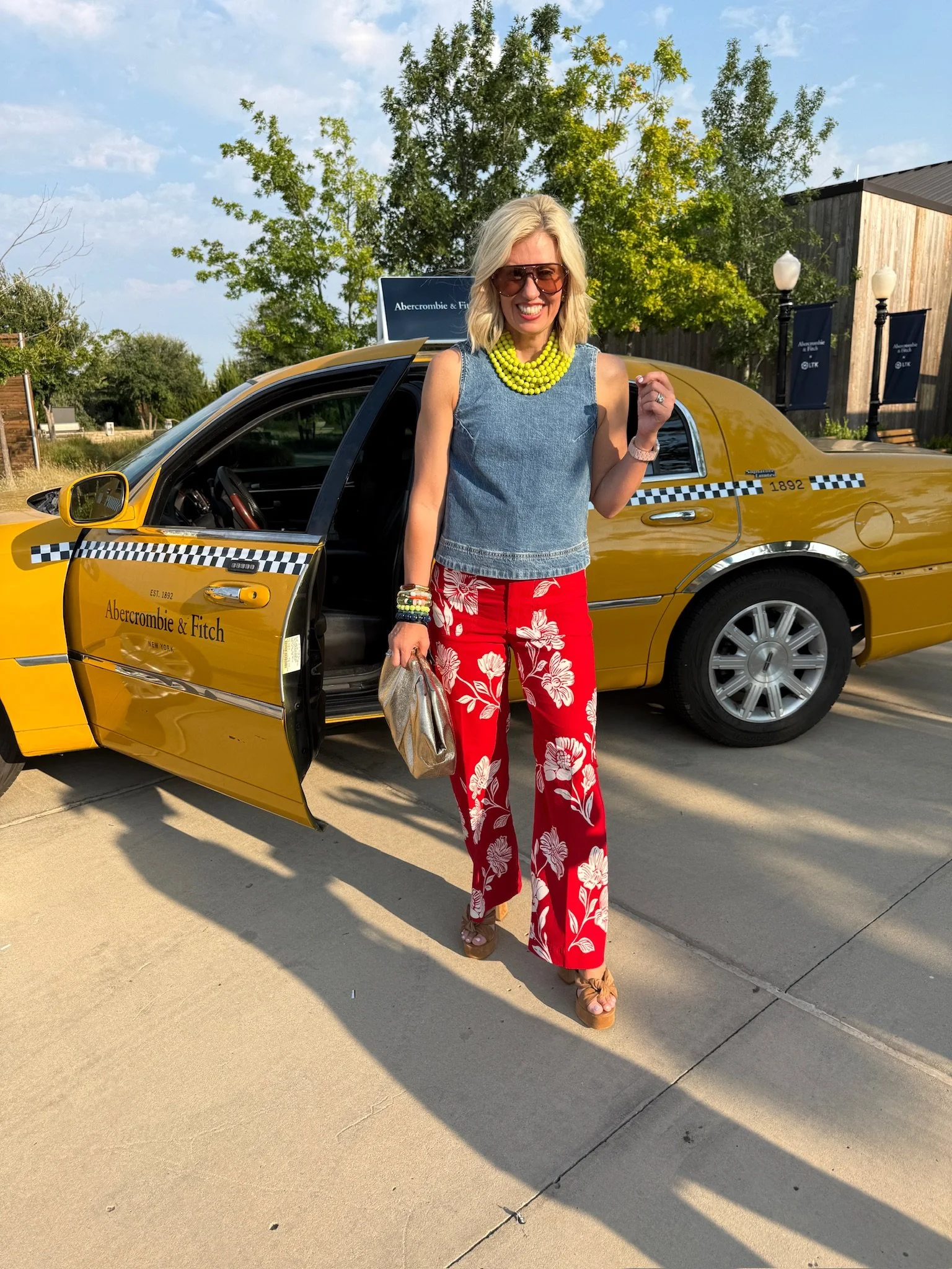 A smiling woman with blonde hair, wearing sunglasses, a sleeveless denim top, red floral pants, and beige platform sandals, standing outside next to a yellow taxi with an Abercrombie & Fitch sign on the roof. She is holding a beige bag and wearing various bracelets and a bright yellow necklace, with a background of green trees, a sky with clouds, and outdoor lamps.