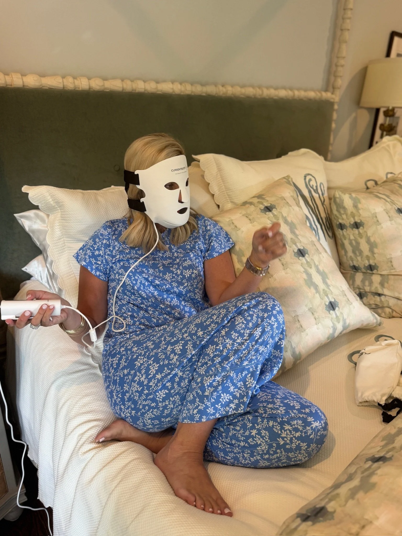 A woman sitting on a bed wearing a white mask and blue floral pajamas, holding a remote control, with a hand touching her ankle.