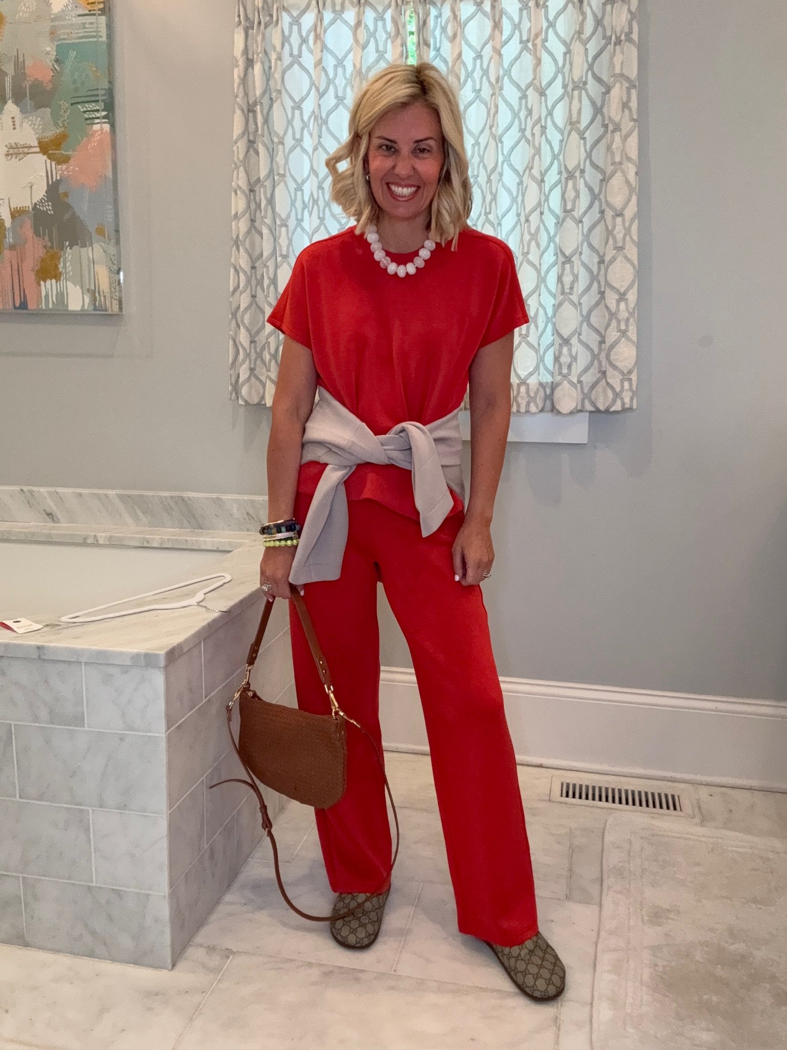 A smiling woman with blonde curly hair standing in a room with light-colored walls and curtains. She is wearing a red outfit, a white beaded necklace, and patterned shoes. She has multiple bracelets on her right wrist and is holding a brown purse with a strap. There is a colorful painting on the wall behind her and a marble-finished surface to her left.