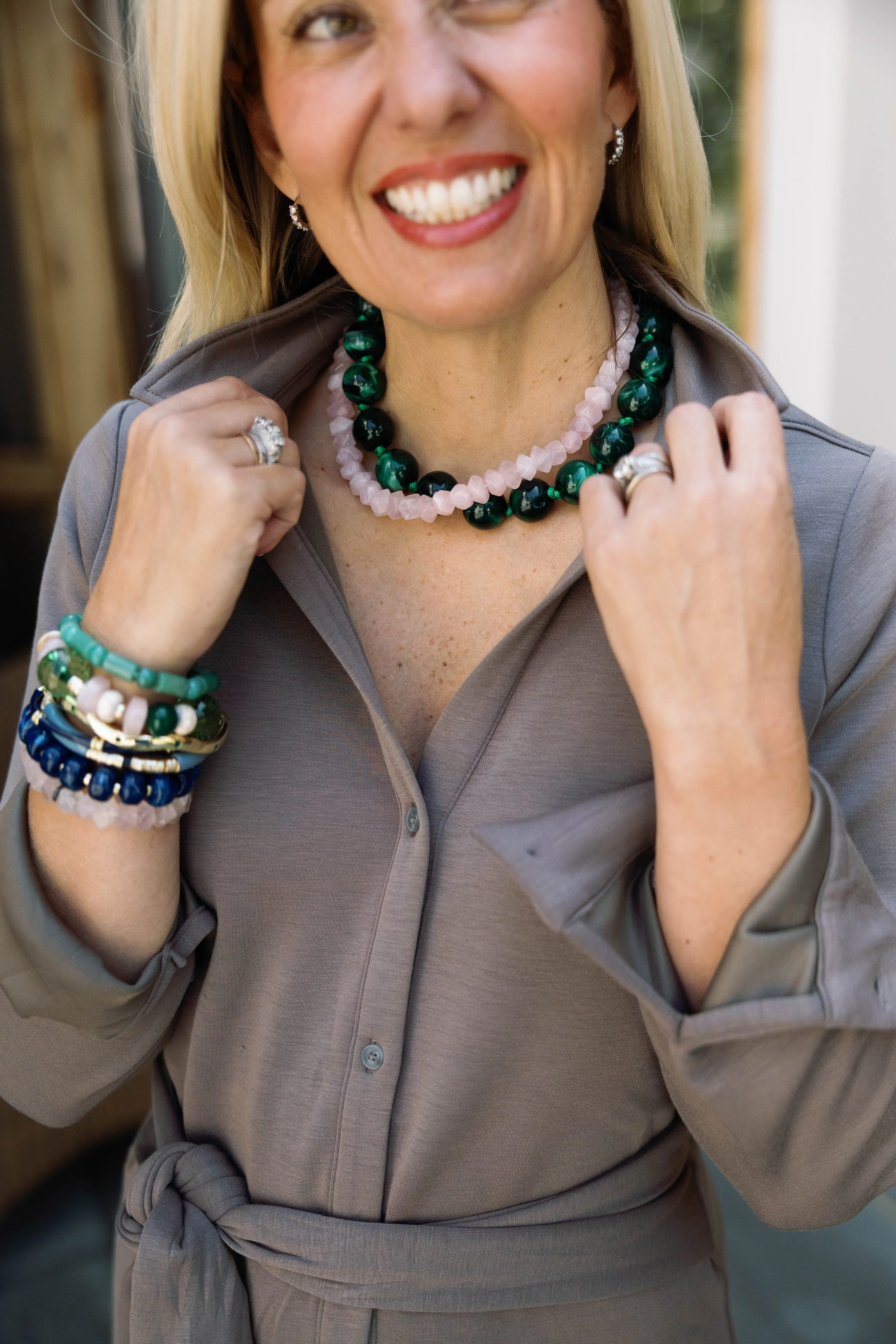 Close-up of a smiling woman wearing jewelry including necklaces, rings, earrings, and bracelets, dressed in a gray collared shirt, with hands touching her collar, in a cheerful and stylish pose.