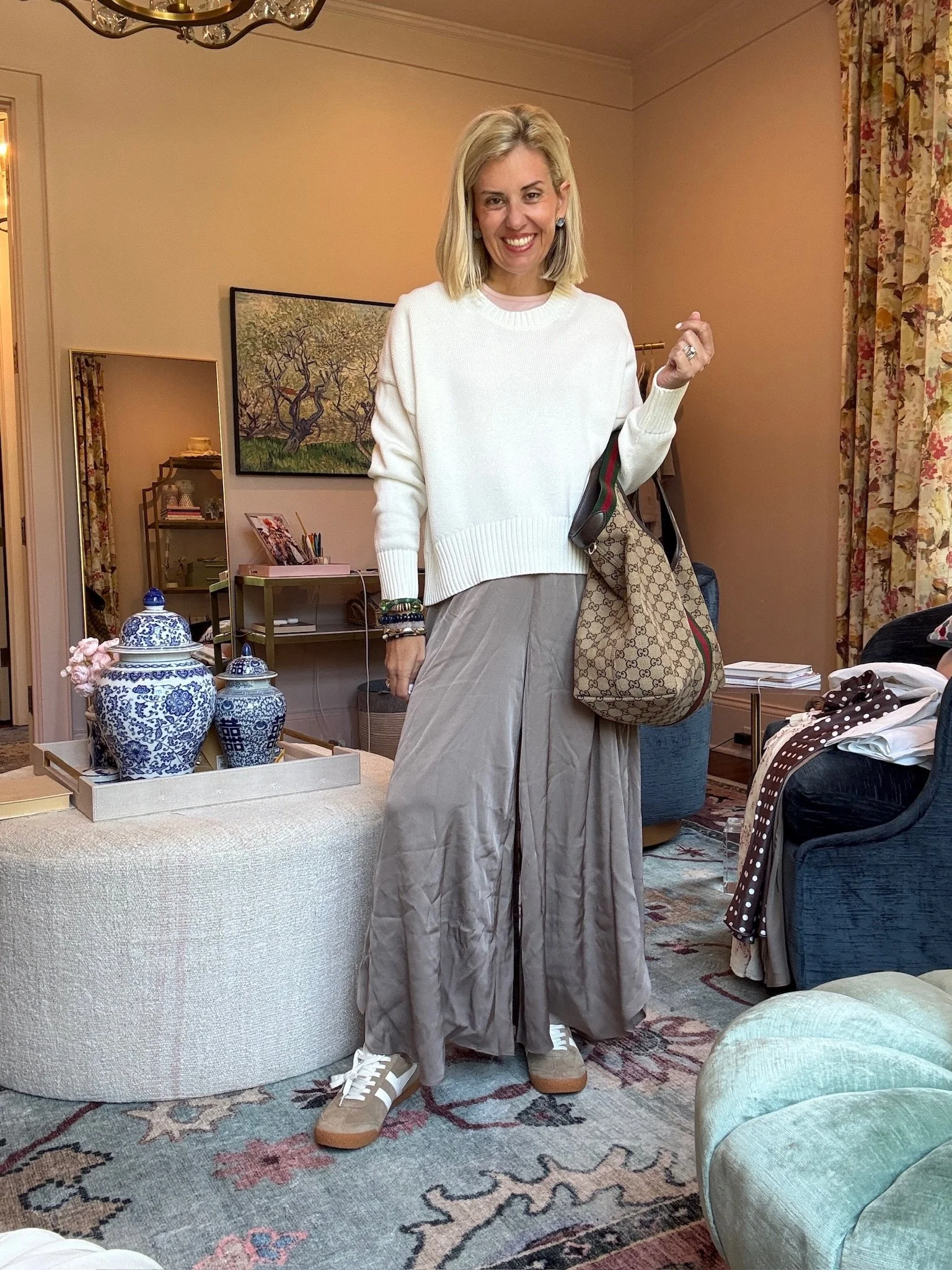 A woman standing in a cozy, well-decorated living room, smiling and holding a Gucci tote bag. She is dressed in wide-leg pants, sneakers, and a white sweater. Behind her, there is a painting of trees, a mirror on a side table, and various decor items, including blue and white vases and a tray with flowers. The room features floral-patterned curtains, a chandelier, and a patterned rug.