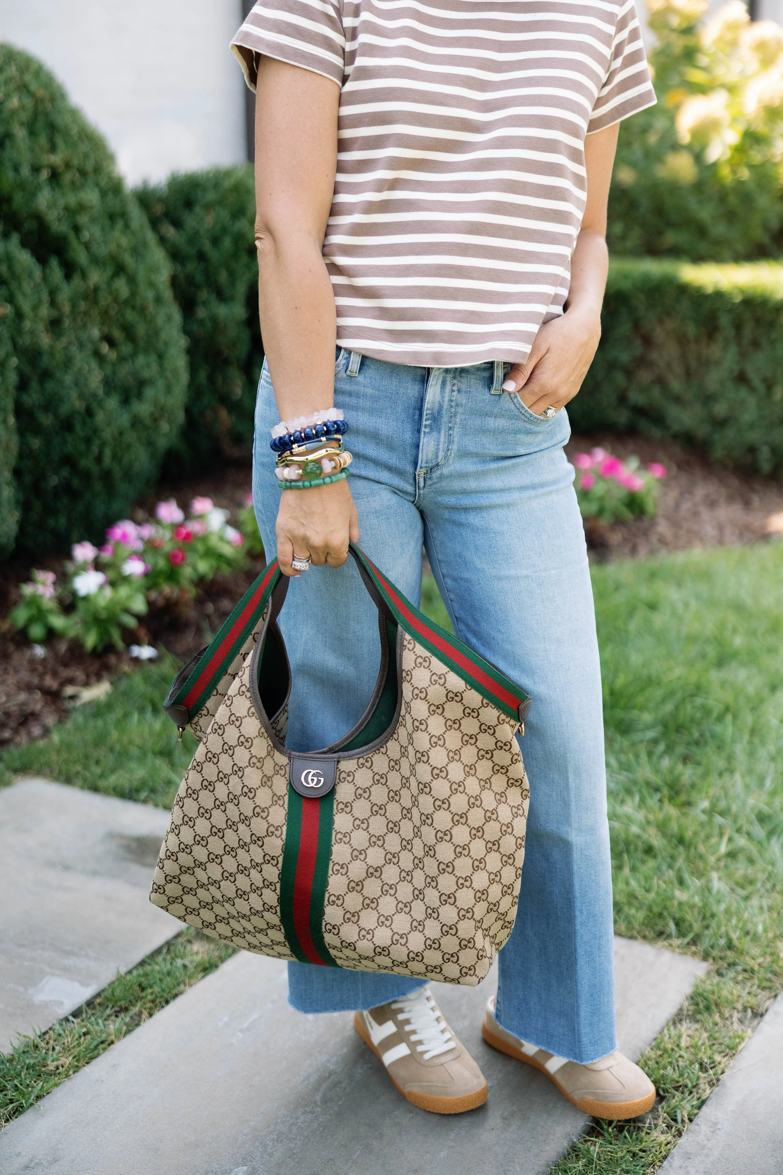 Person wearing a brown and white striped T-shirt, blue jeans, and beige and white sneakers, holding a Gucci tote bag with the GG pattern and green-red-green straps, standing on a sidewalk with a garden in the background.
