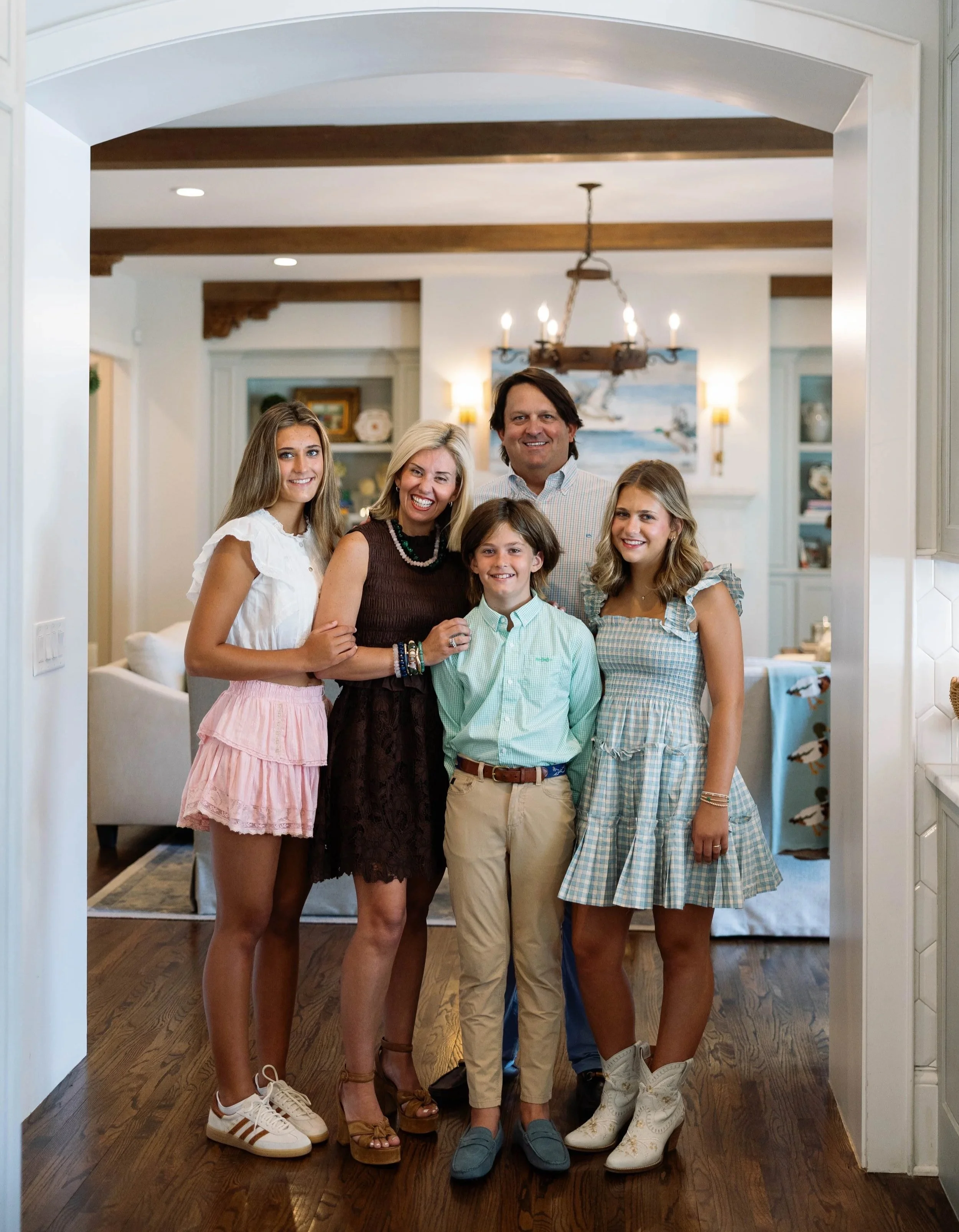 A family of six posing together in a home. The group includes three women, two girls, and one man. They are standing on a wooden floor in front of a white archway, with a background of bookshelves, paintings, and warm lighting.