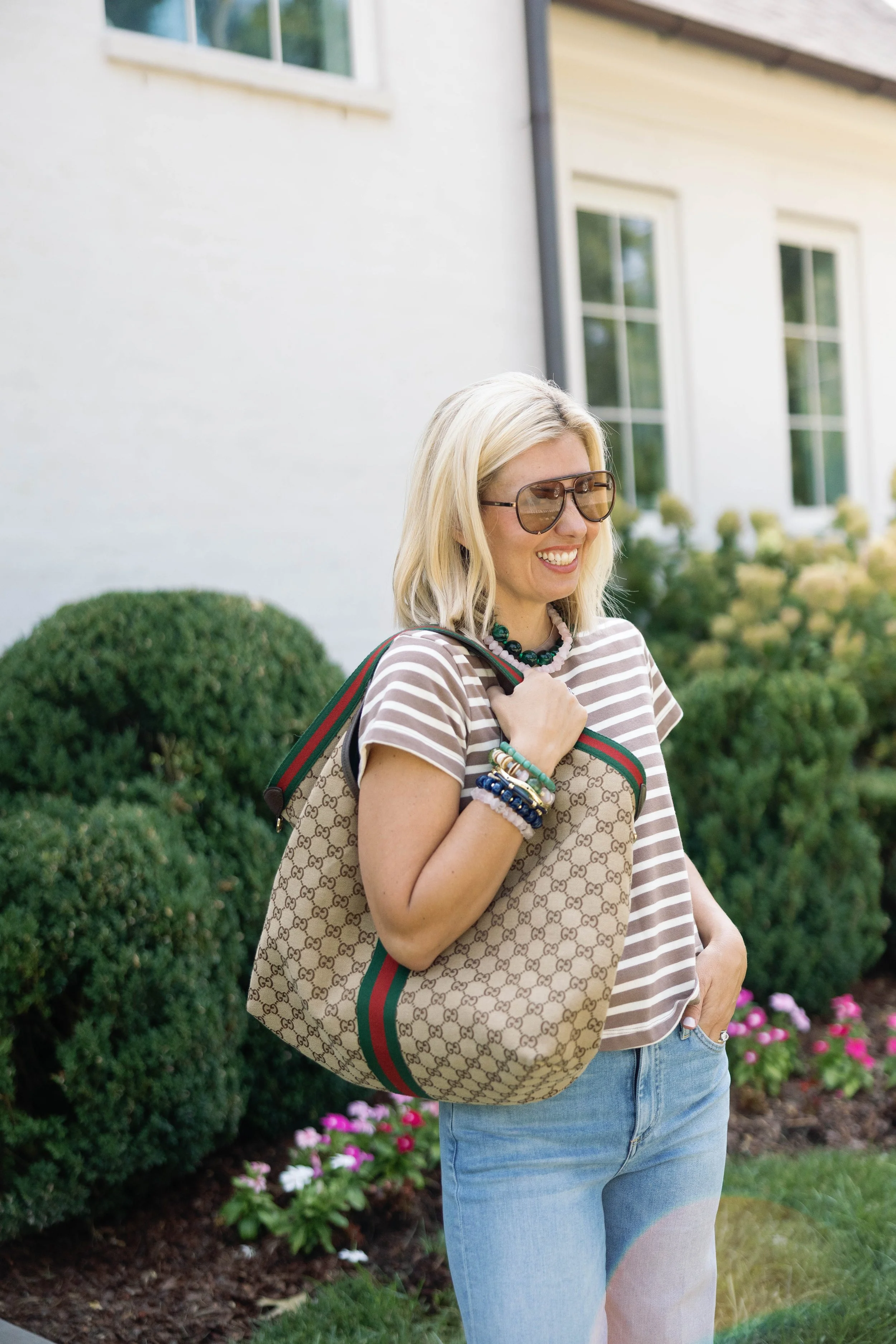 A woman with blonde hair, wearing sunglasses, a striped shirt, and jeans, smiling and carrying a designer handbag with a pattern and a red and green striped strap, standing outdoors in front of a house with white walls and large windows, surrounded by flowering plants and bushes.