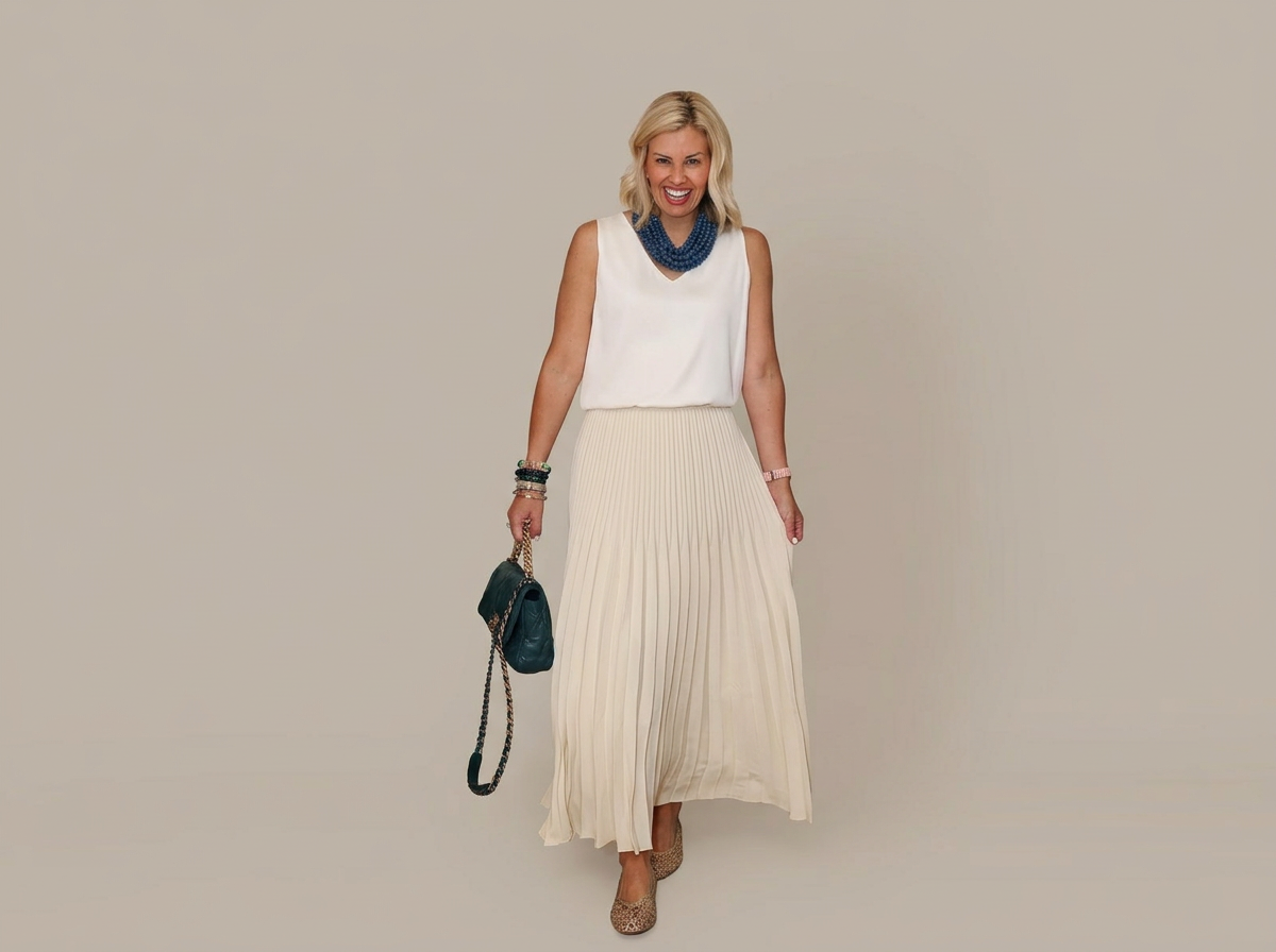 A woman smiling and walking, wearing a white sleeveless top, a long pleated ivory skirt, a blue necklace, and holding a black purse with a chain strap. She has blonde hair and is wearing nude heels.