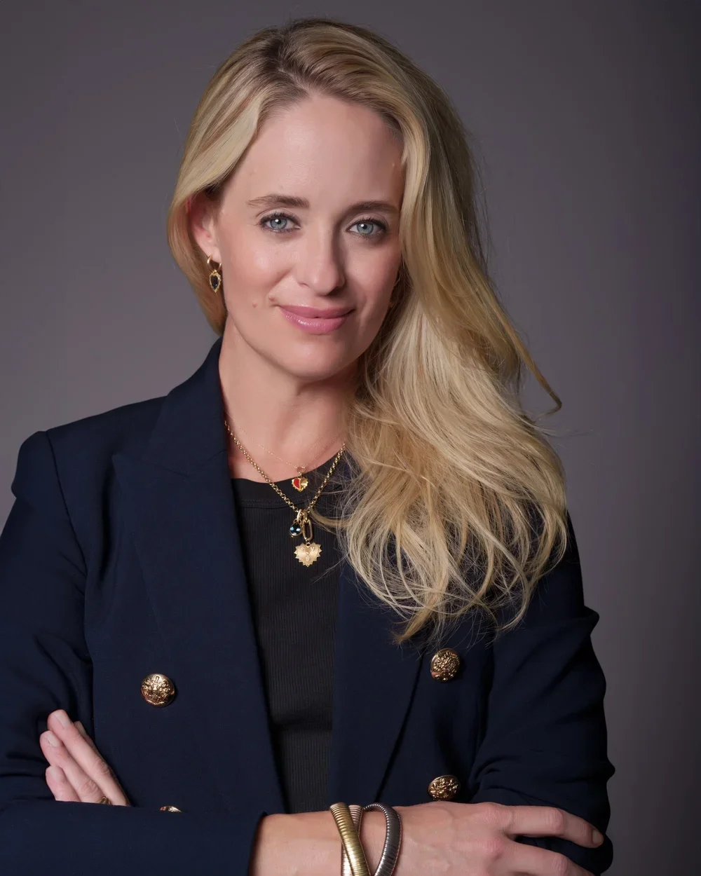 A woman with long blond hair and blue eyes, wearing a navy blazer with gold buttons, gold jewelry, and a black top, standing against a gray background.