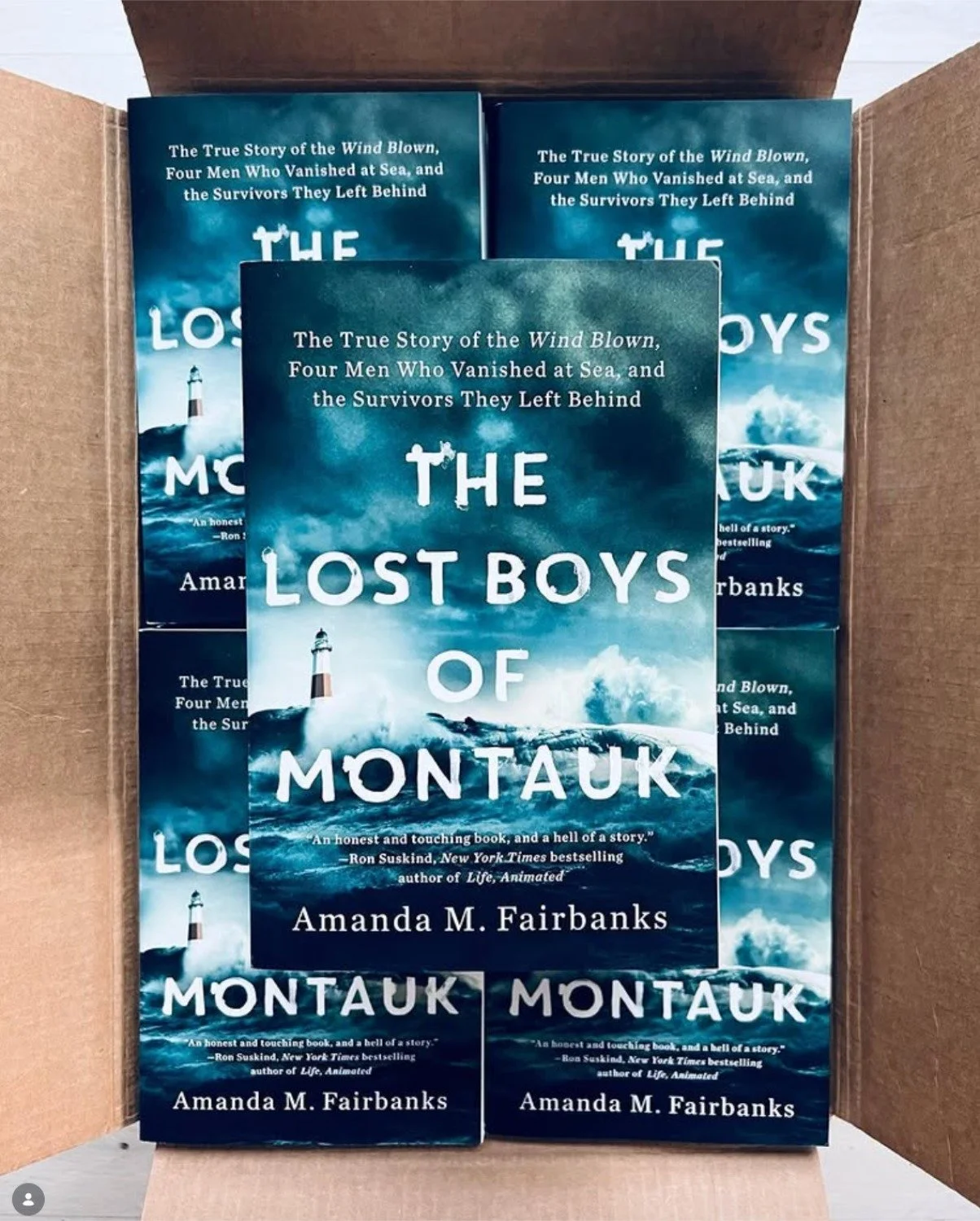 Three copies of a book titled 'The Lost Boys of Montauk' by Amanda M. Fairbanks inside a cardboard box. The book cover features a stormy sea with a lighthouse and waves.