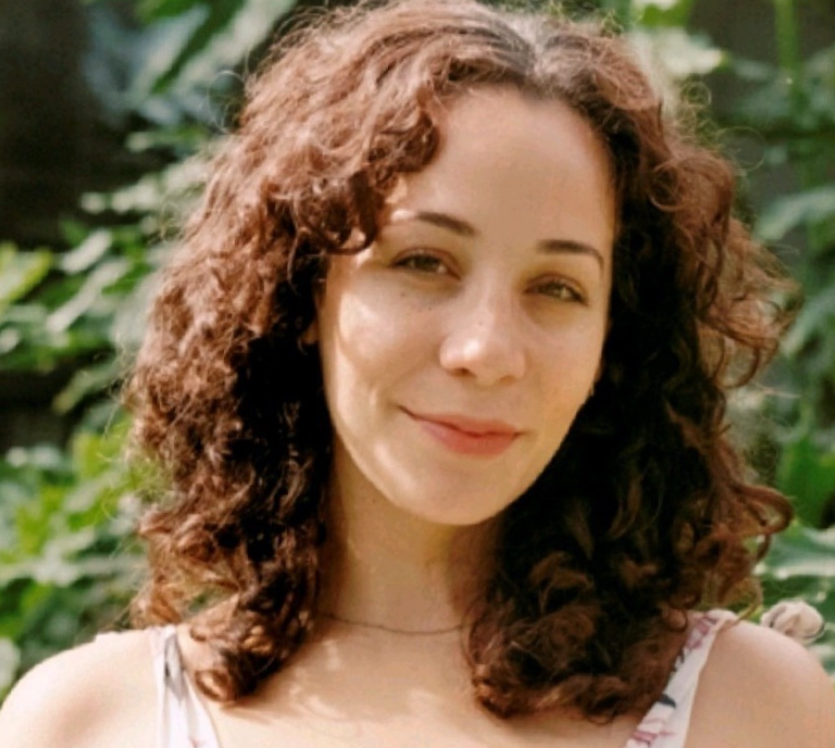 A woman with curly brown hair smiling outdoors with green foliage in the background.