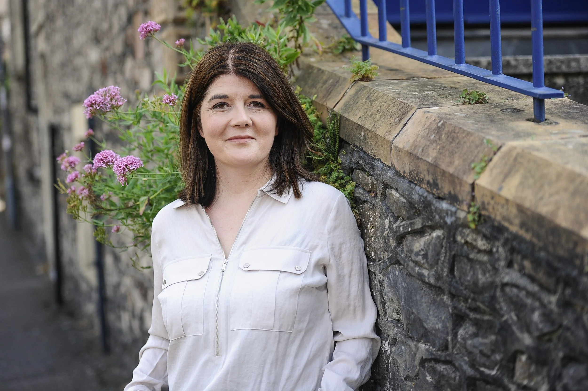 A woman with shoulder-length dark brown hair and light skin, wearing a white zip-up jacket, leaning against a stone wall with pink flowers and green plants in the background.