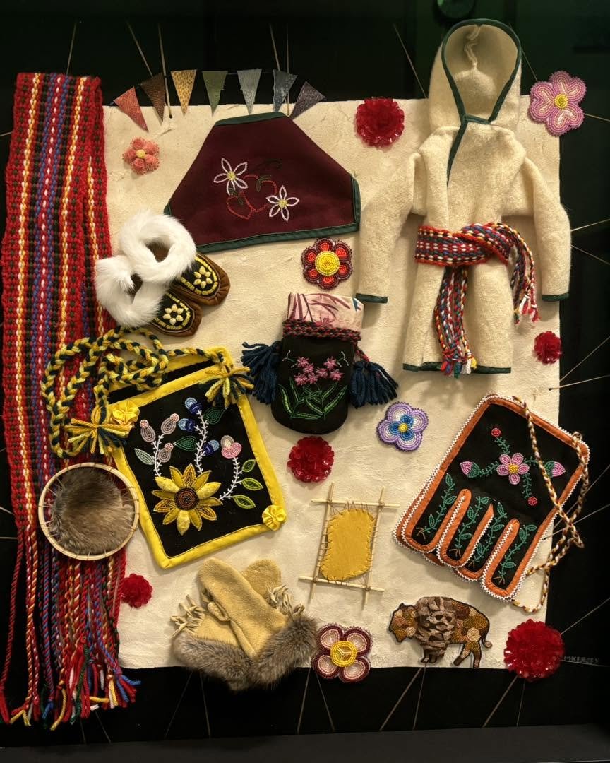 A collection of traditional and handmade items, including embroidered bags, moccasins, beadwork, tufted pieces, and woven accessories, displayed on Elk hide, backed with black Melton Wool.