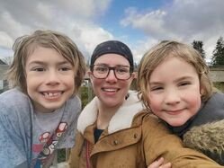 Three smiling people outdoors, two young boys and an adult woman, with a cloudy sky and trees in the background.