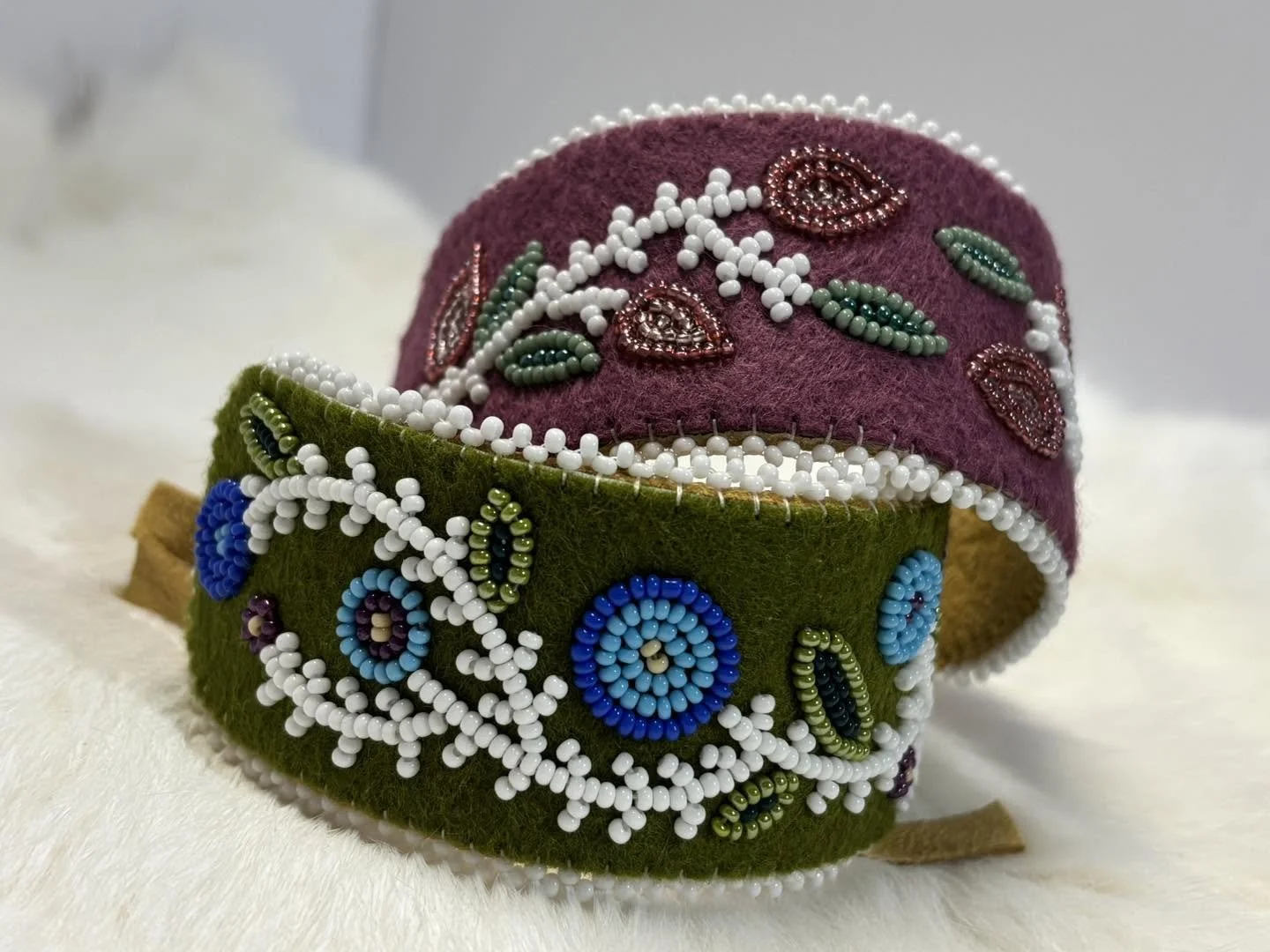 Two beaded cuff bracelets, one purple with white, green, and brown bead patterns, and one green with blue, purple, green, and white bead patterns, placed on a white fluffy surface.
