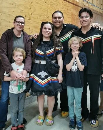 Mara, her children, and the Ivan Flett Memorial Dancers standing together indoors against a wooden wall, smiling at the camera.