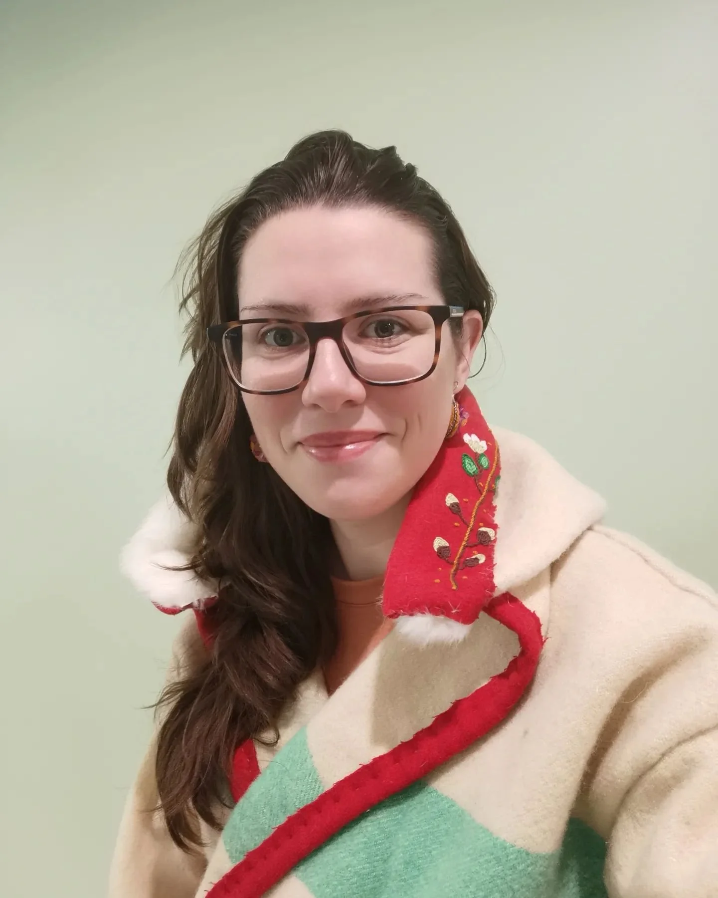 A woman with long brown hair and glasses smiling at the camera, wearing a hand-sewn Capote with a red and green design, standing against a plain light green wall.