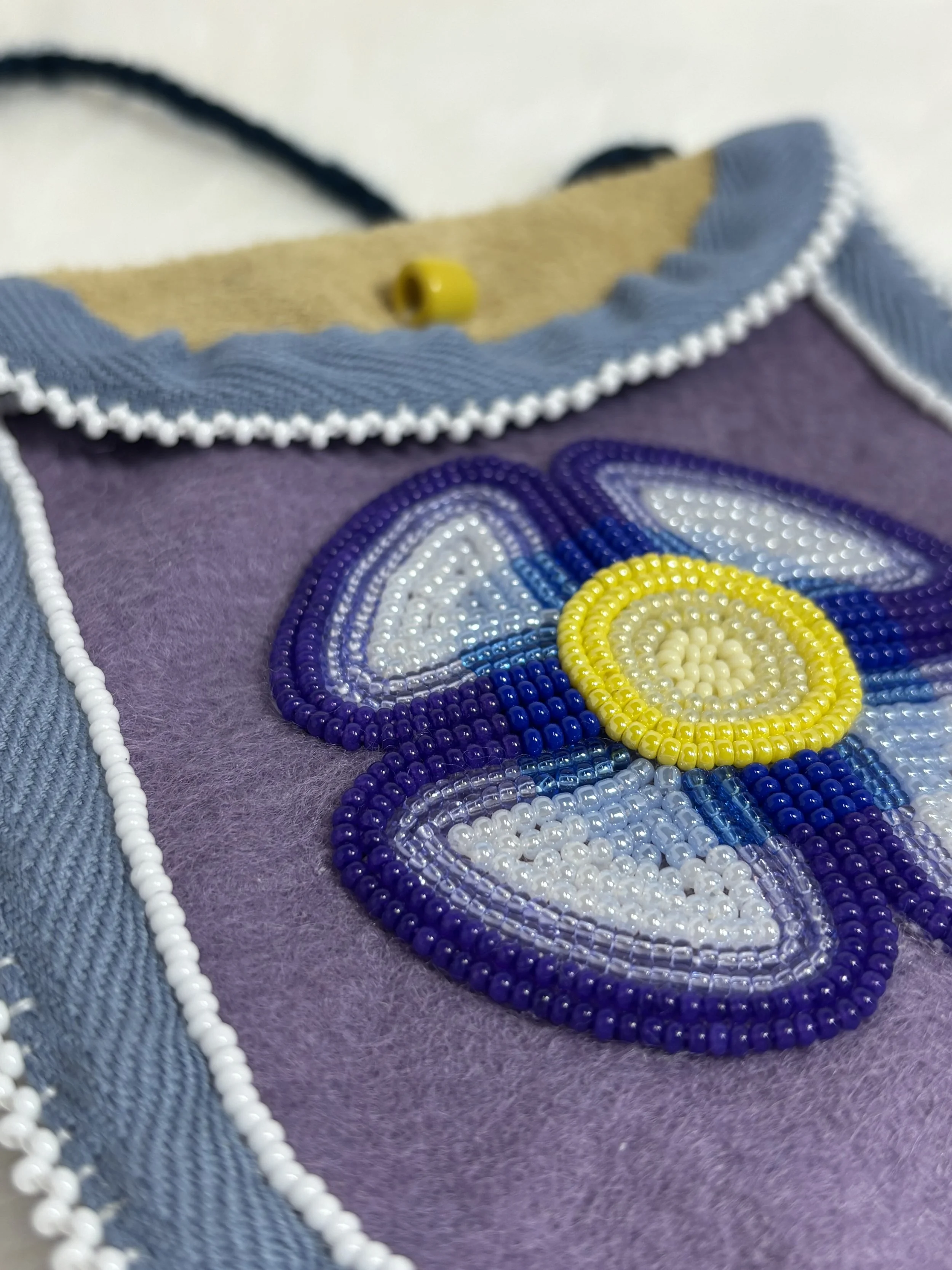 Close-up of colorful beadwork on a medicine bag, with purple, yellow, white, and blue beads forming a floral pattern, on purple wool and bordered by blue and white trim.