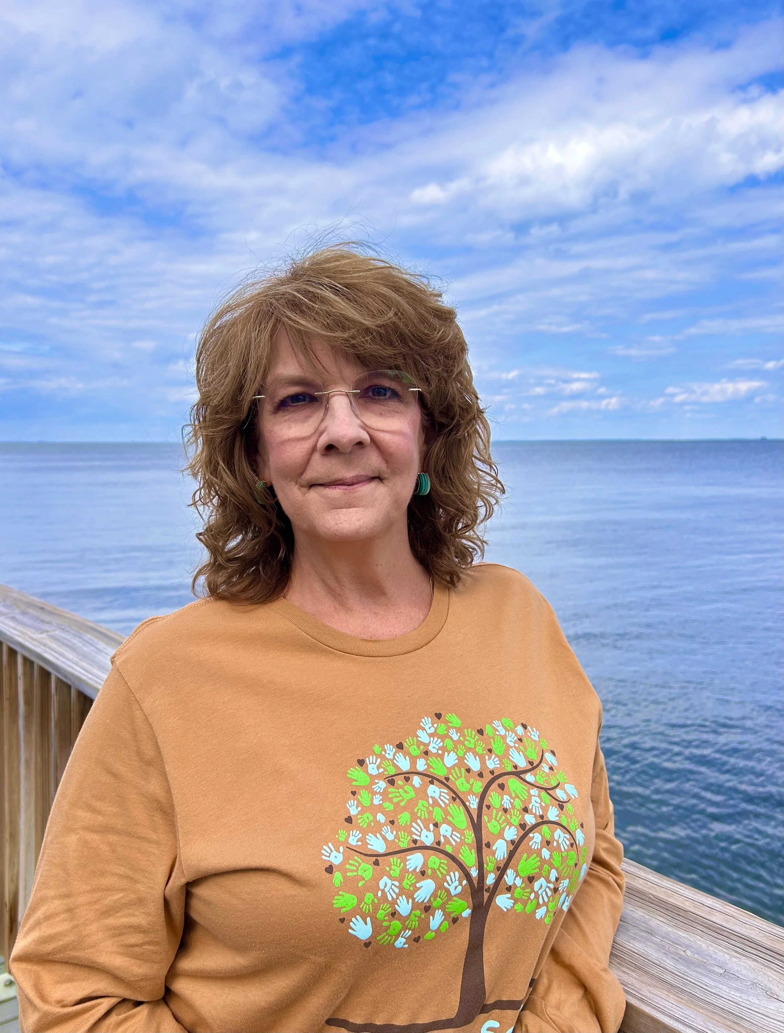 A woman with glasses and wavy auburn hair standing on a wooden boardwalk near water, wearing a tan sweatshirt with a colorful tree of life design, against a background of a blue sky with clouds.