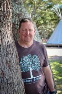 A man smiling outdoors next to a large tree, wearing a dark T-shirt with a tree graphic and words 'Future School'.