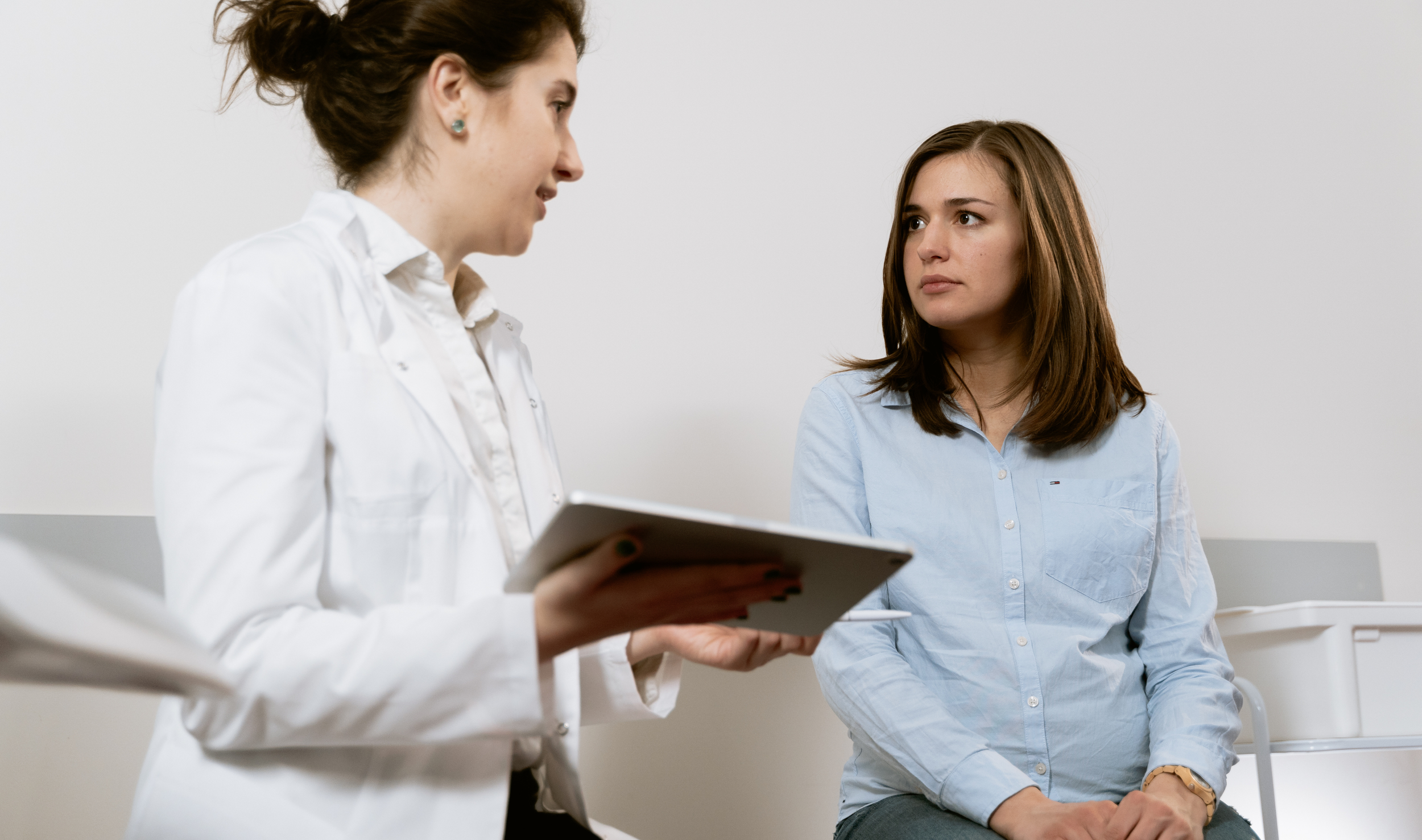 Two women having a conversation in a clinical or office setting. One woman is holding a tablet and the other is listening attentively. Both are dressed casually, with neutral background.