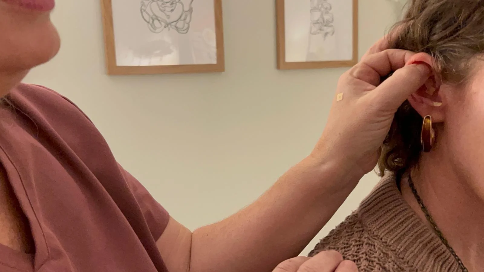 A person is helping another person to put on an ear cuff, with one hand holding the ear and the other holding the cuff. The background has framed line drawings on the wall.