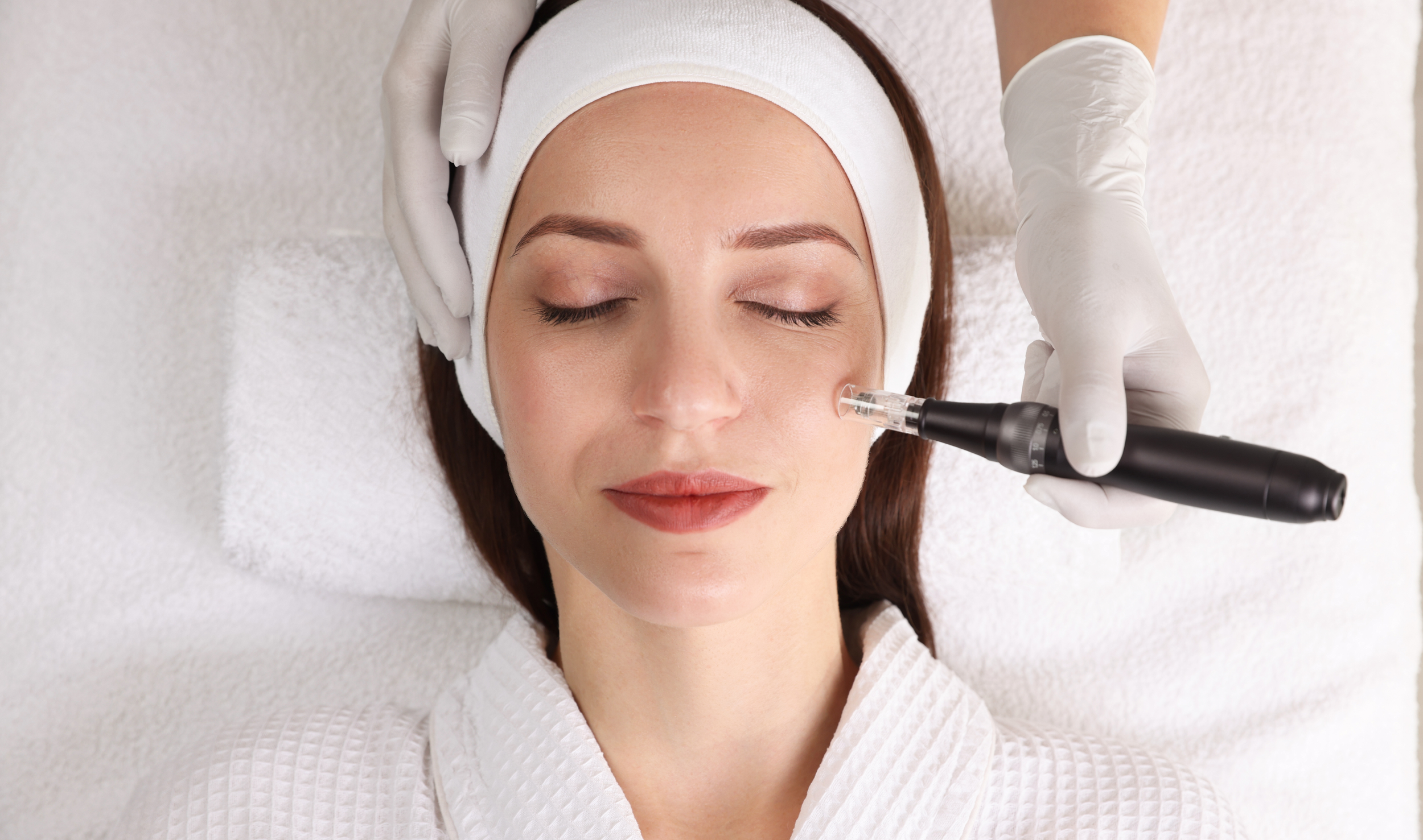 A woman lying down with eyes closed, receiving a facial treatment with a procedure device near her cheek, wearing a white headband and a white robe, in a spa or cosmetic clinic setting.