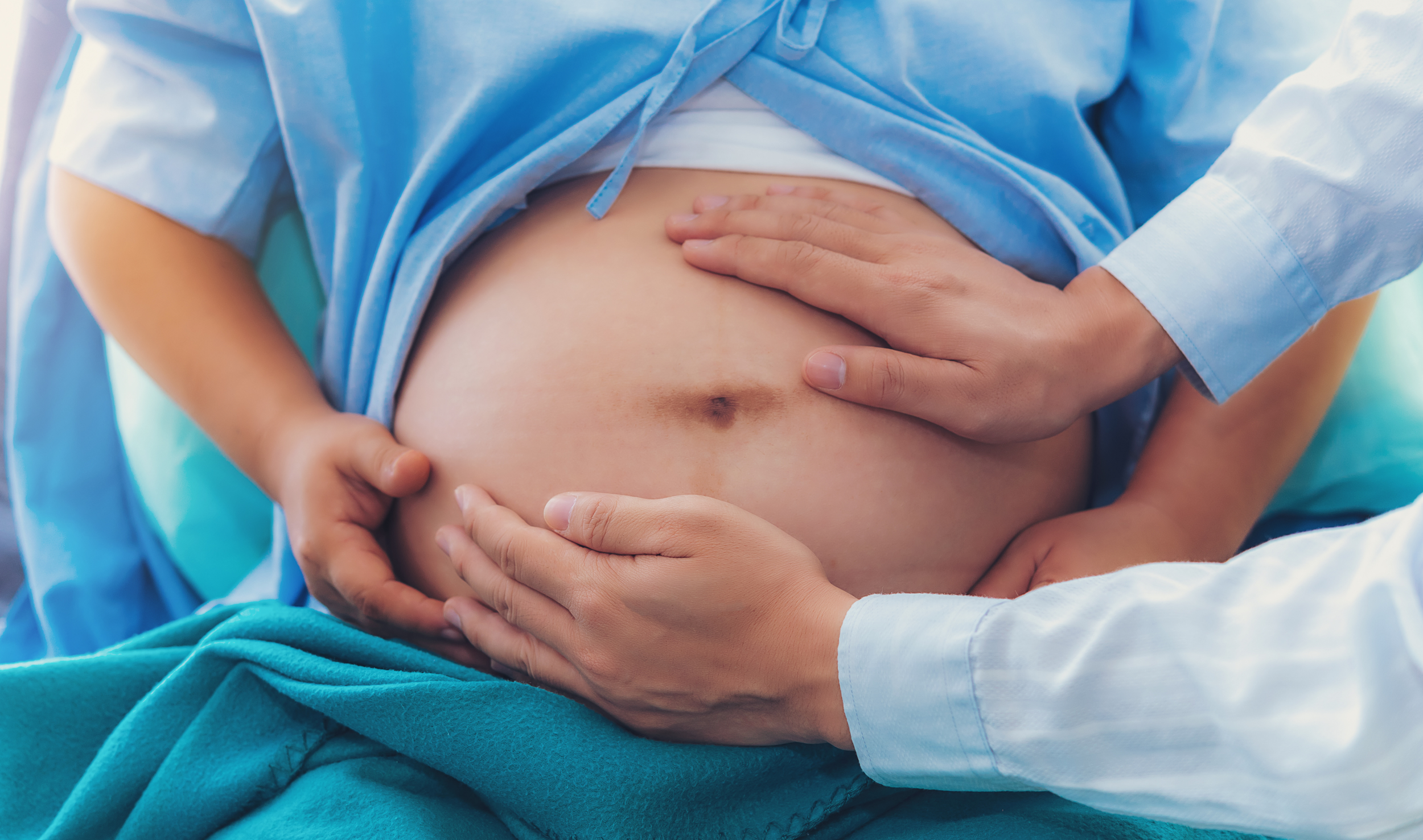 Close-up of a pregnant woman's belly with hands cradling it, wearing a blue hospital gown and a white shirt underneath.