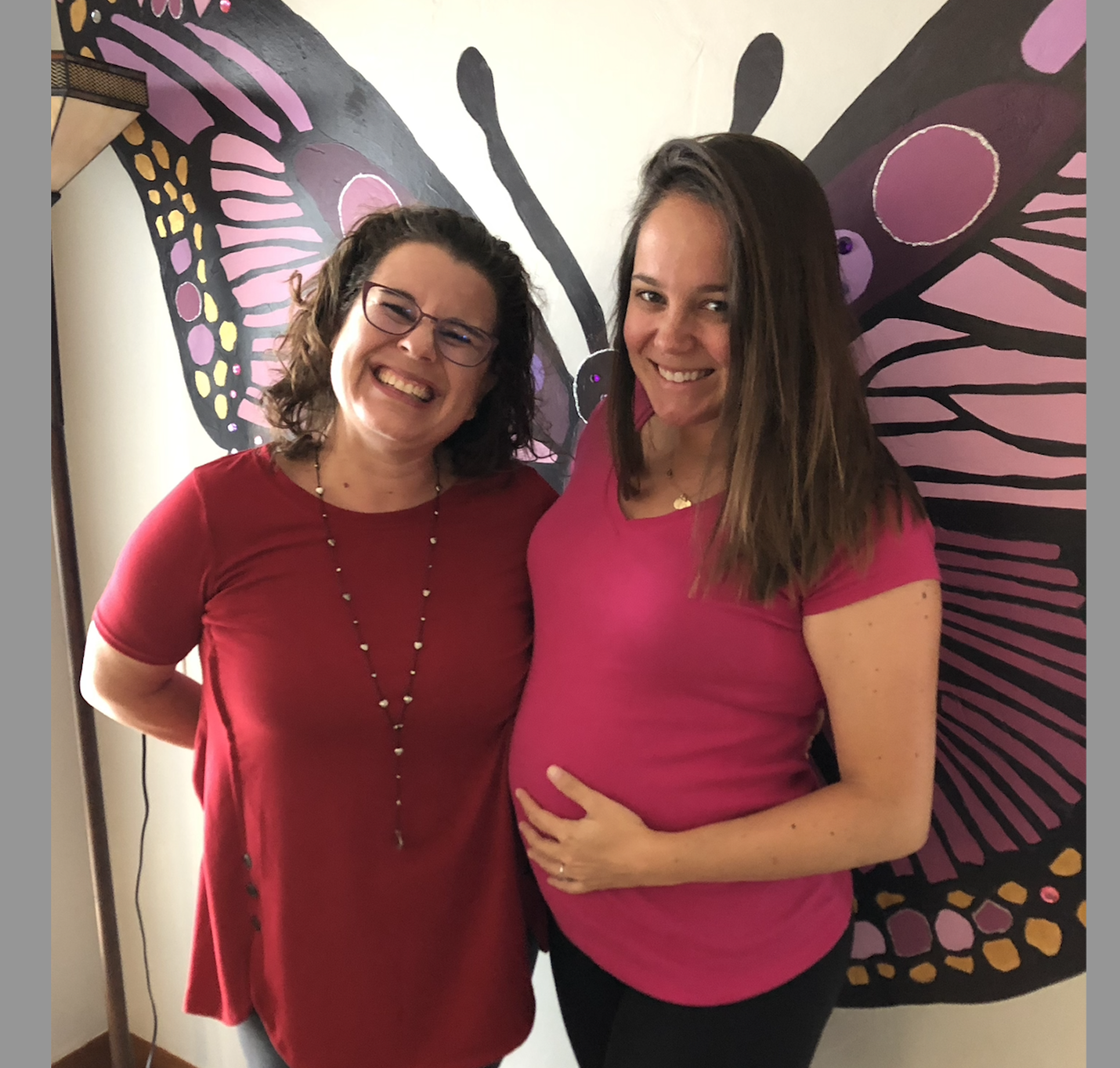 Two women smiling, one with glasses and wearing a red shirt, the other pregnant with long hair, holding her belly and wearing a pink shirt, standing in front of a large butterfly wall art.
