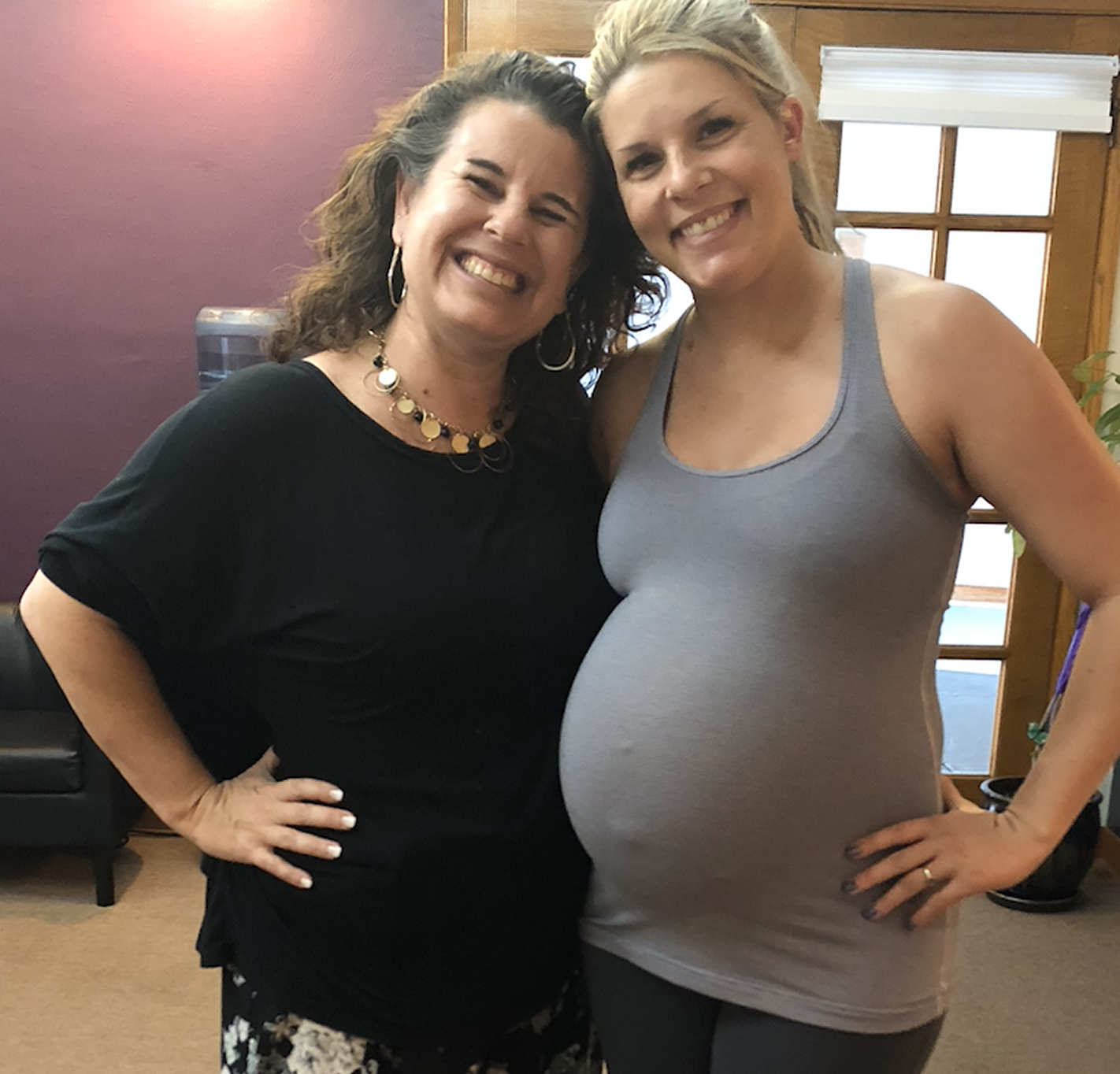 Two women smiling and posing together indoors, one wearing a black top and the other wearing a gray tank top, with one woman visibly pregnant.