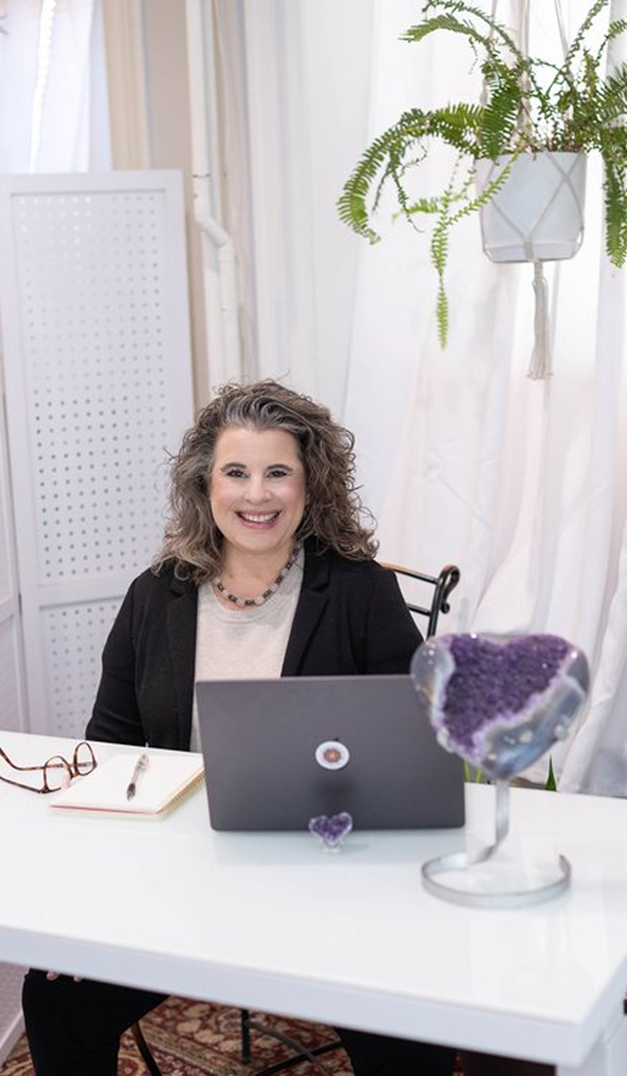 A woman with curly gray hair smiling, sitting at a white desk with a laptop, notebook, and glasses. Behind her is a white folding room divider, and a potted fern in a white hanging planter. In the foreground, there is a purple geode display on a stand.