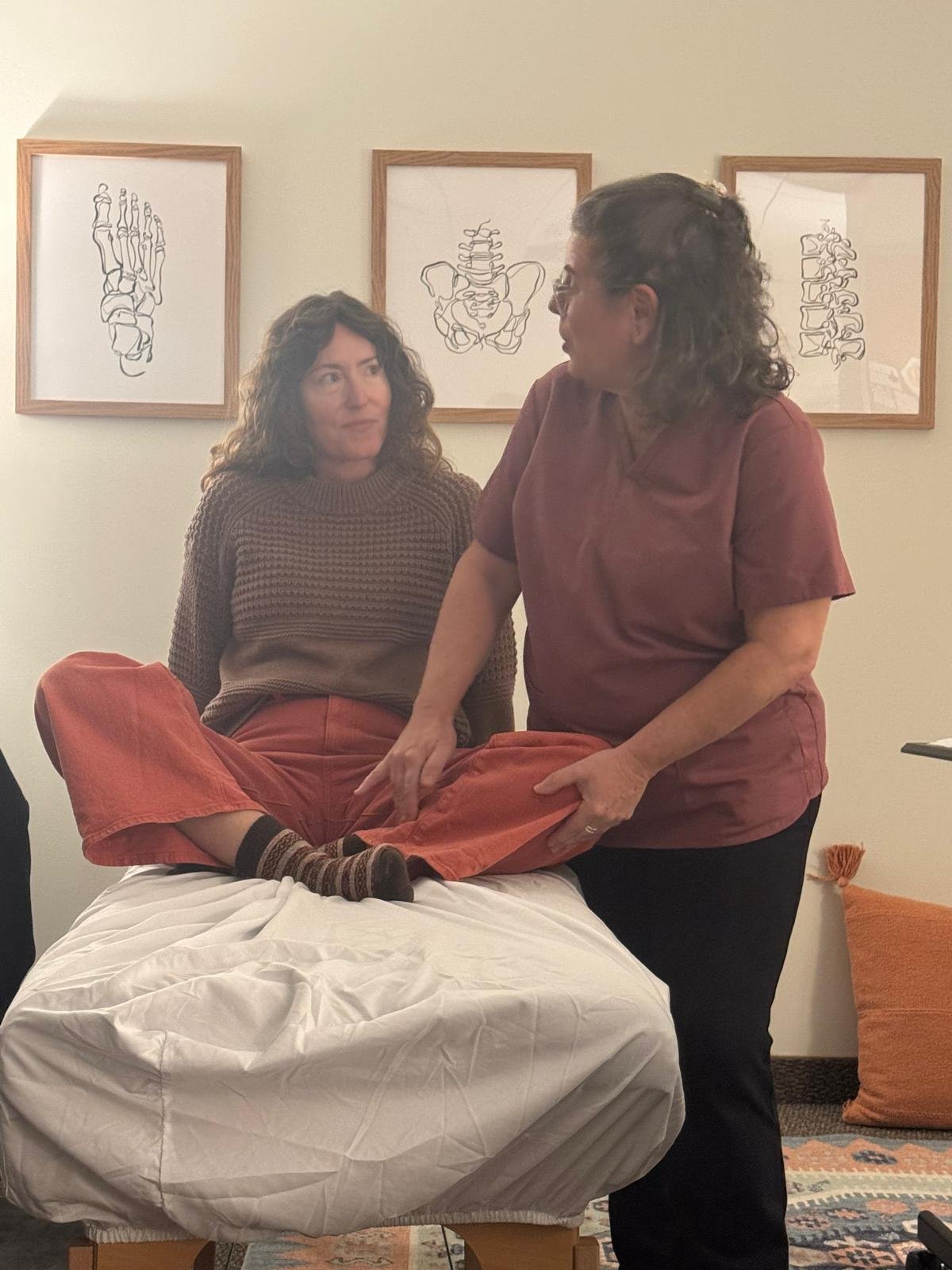 Two women in a room with framed anatomical line drawings on the wall, one sitting cross-legged on a padded table or massage table, the other standing beside her, holding her foot and appearing to give a massage or therapy.