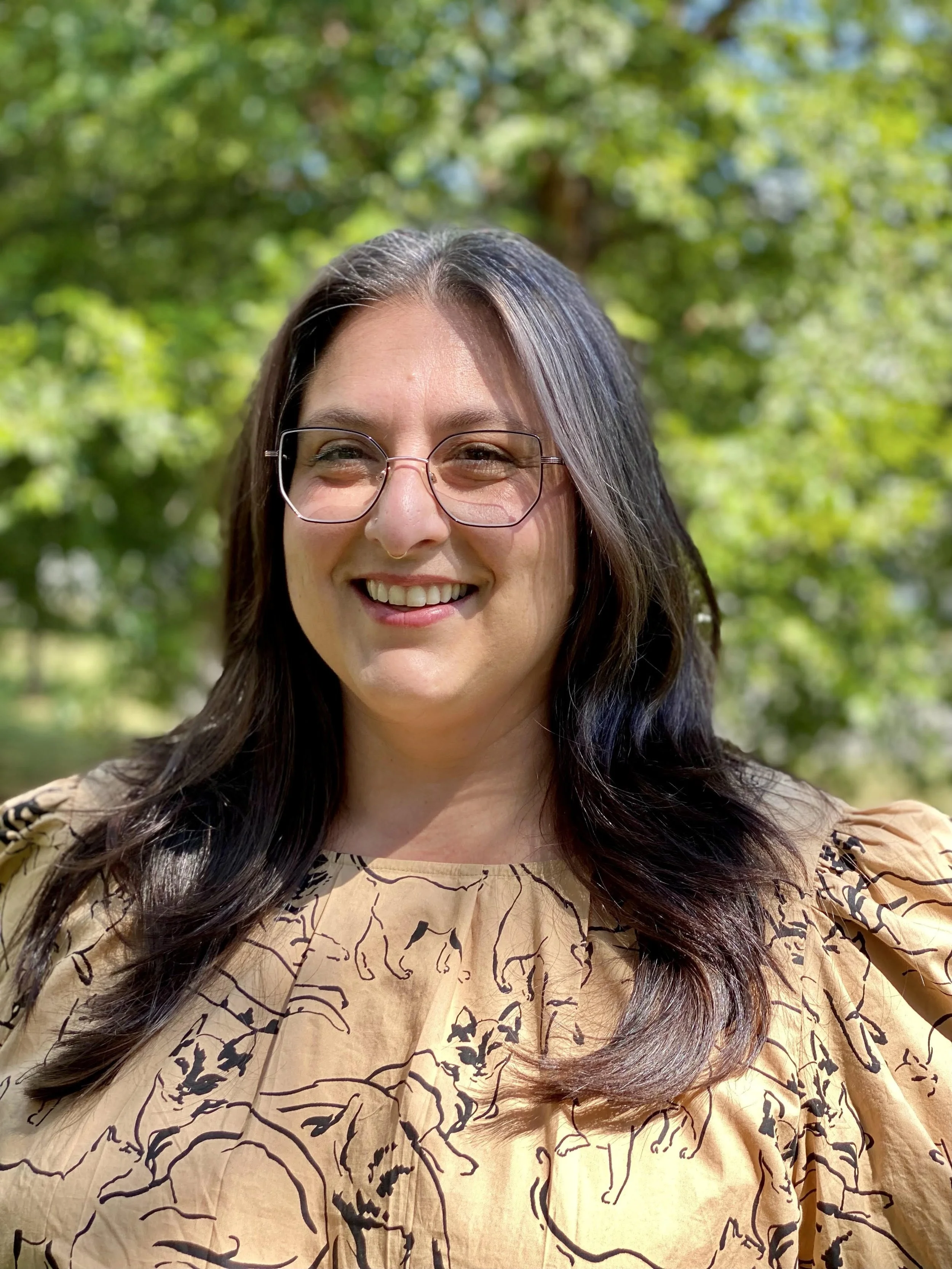 A woman with long dark hair, wearing glasses, smiling, and dressed in a beige top with black abstract patterns, standing outdoors with a blurred background of green trees.