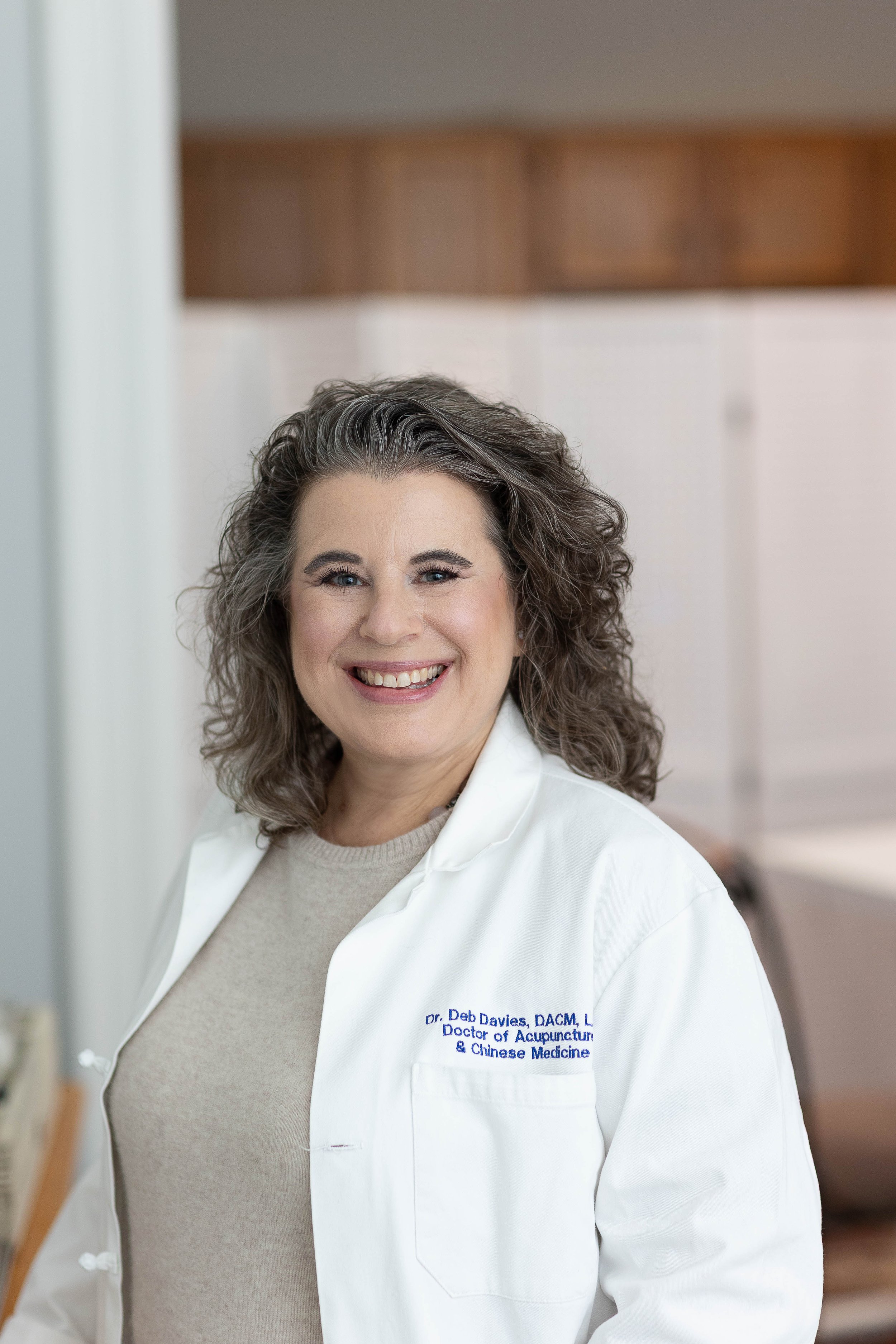 A smiling woman with curly gray hair wearing a white medical coat with embroidery that reads Dr. Deb Davies, DACM, Doctor of Acupuncture & Chinese Medicine, standing in a clinic or medical office.
