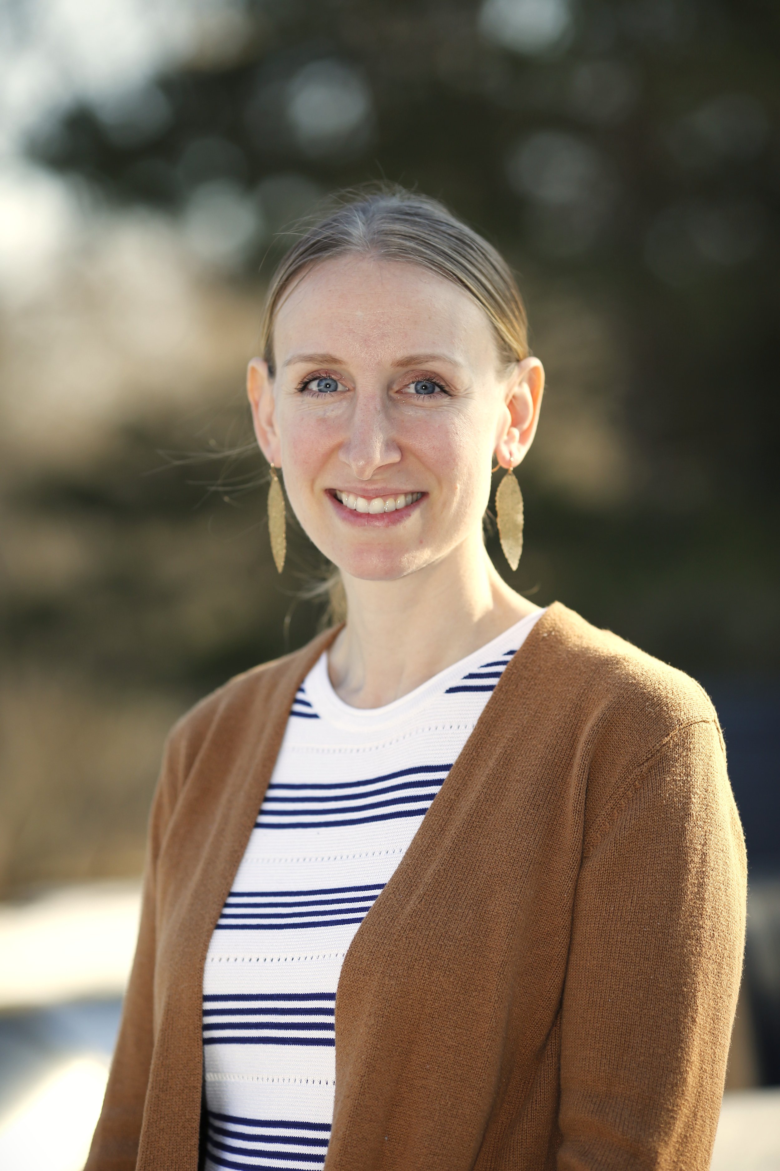 A woman with blue eyes, light brown hair, and earrings outdoors, smiling and wearing a striped shirt and a brown cardigan.