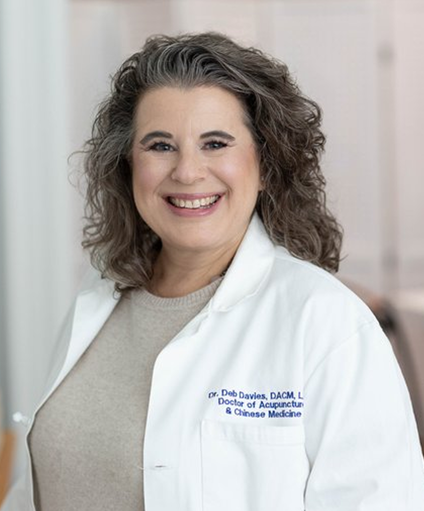 A smiling woman with curly hair, wearing a white lab coat with her name and credentials embroidered on it, standing in a professional medical setting.