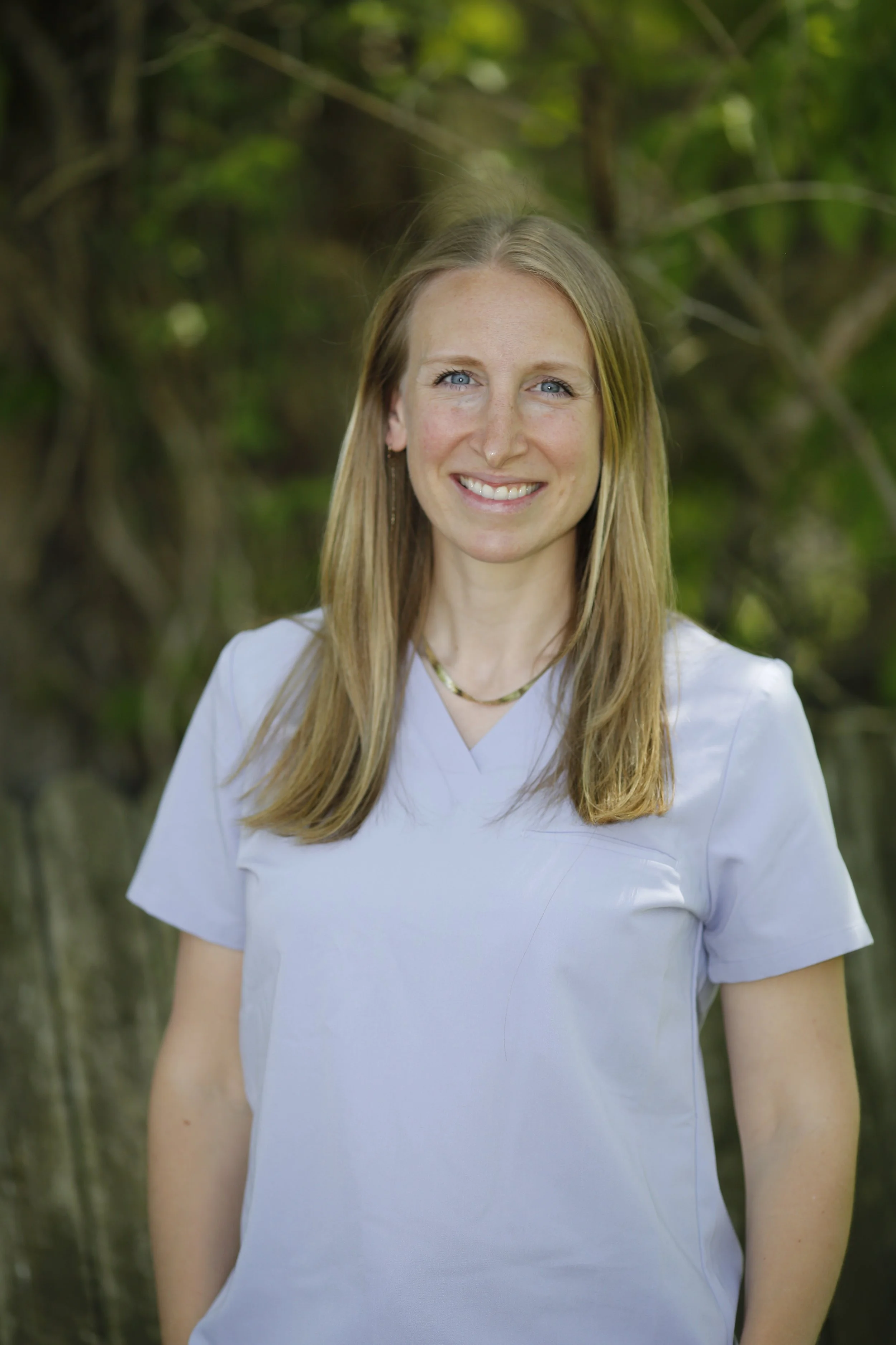 A woman with long blonde hair, blue eyes, and a white shirt, smiling outdoors with a background of trees and green foliage.