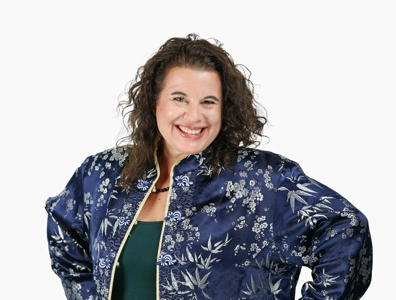 A woman with curly brown hair smiling, wearing a dark blue satin jacket with floral print and a gold zipper, over a green top.