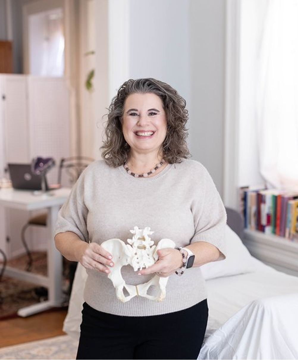 A smiling woman with curly gray hair holds a model of a human pelvis in a well-lit room with a bed, shelves of books, and a desk in the background.