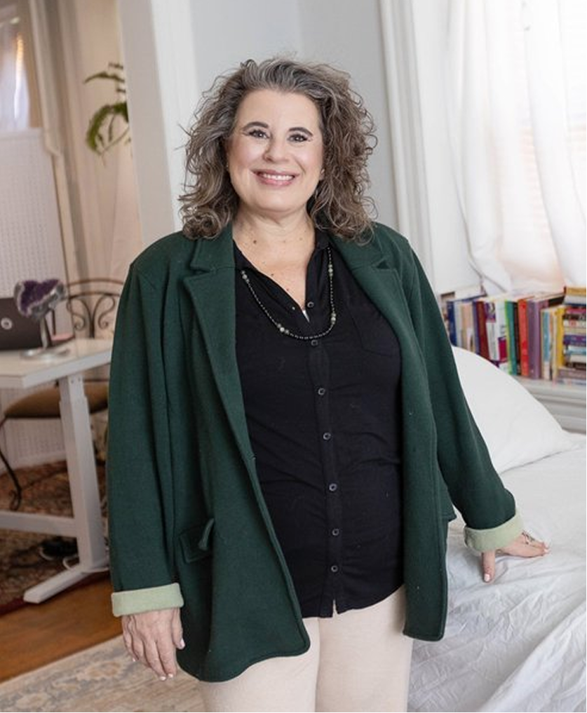 A woman with curly gray hair smiling, wearing a black top and dark green blazer, standing in a cozy room with a bed, bookshelf, and curtains.