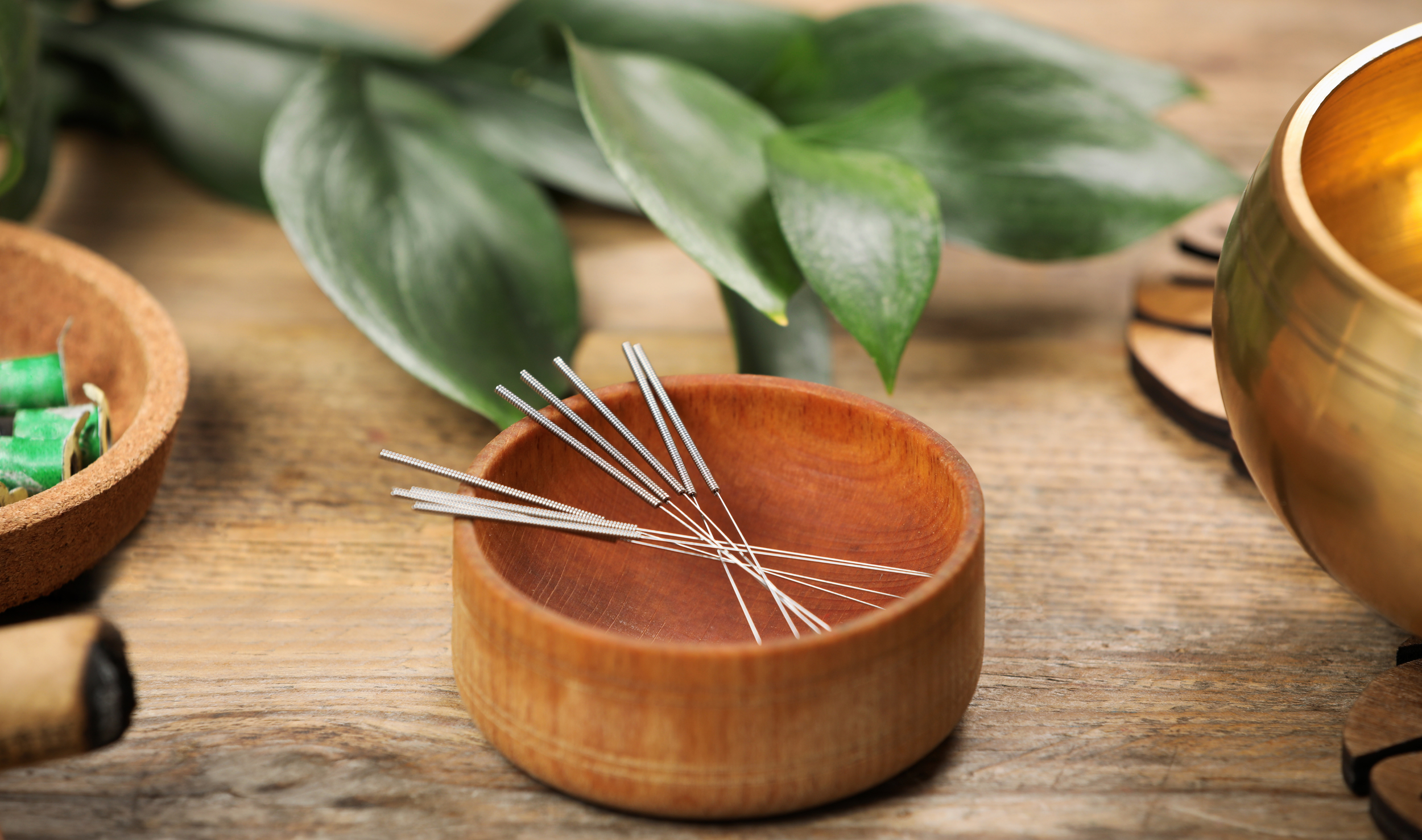 A wooden table with acupuncture needles in a small round wooden bowl, surrounded by green leaves, a golden cup, and other items.