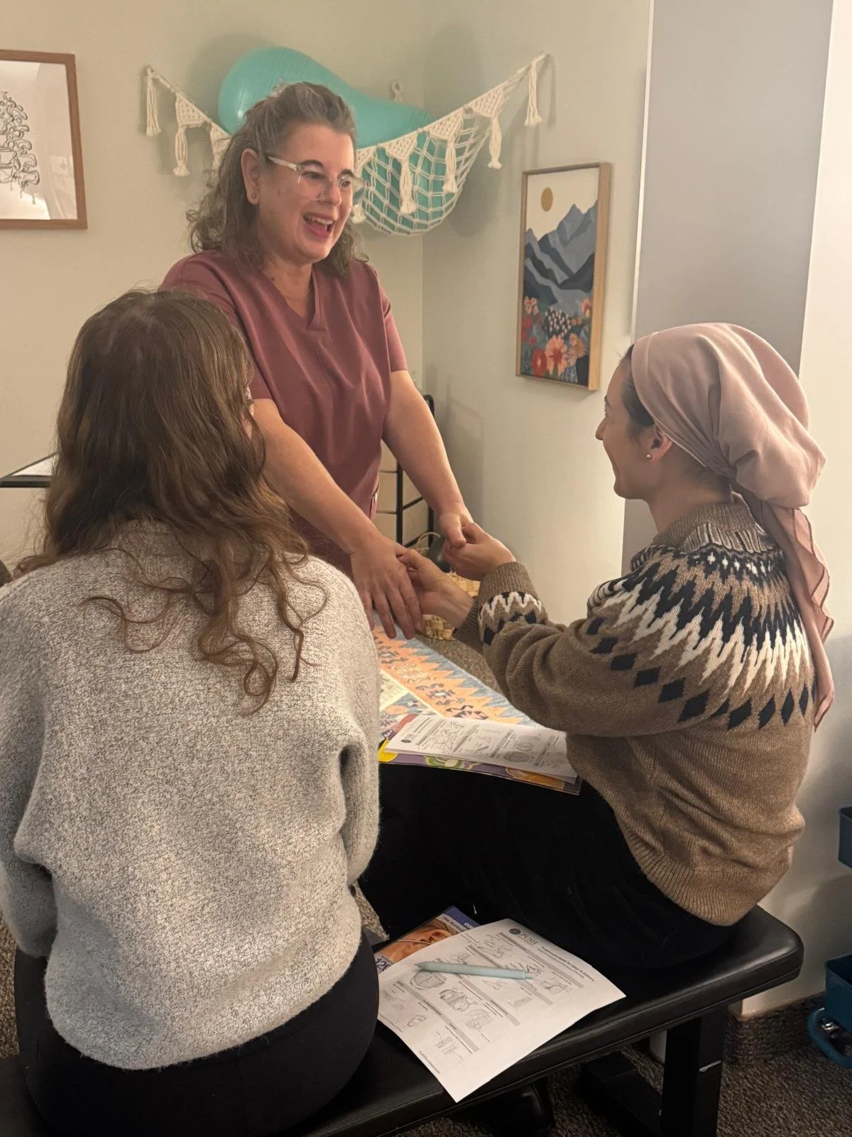 Three women interacting in a cozy room, with one woman smiling and holding hands of another woman, while the third woman is sitting with her back to the camera, wearing a light gray sweater and black pants.