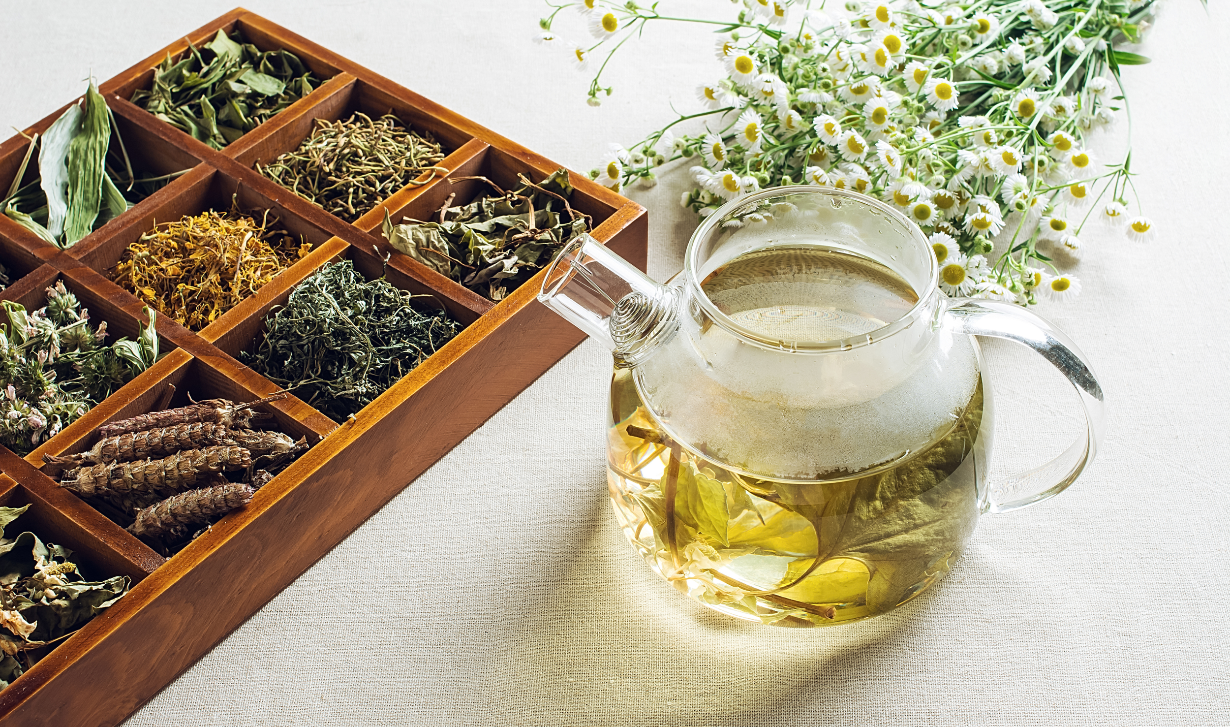 A wooden box with twelve compartments filled with different dried herbs and flowers, a glass teapot with herbal tea, and a bunch of fresh daisies on a white tablecloth.