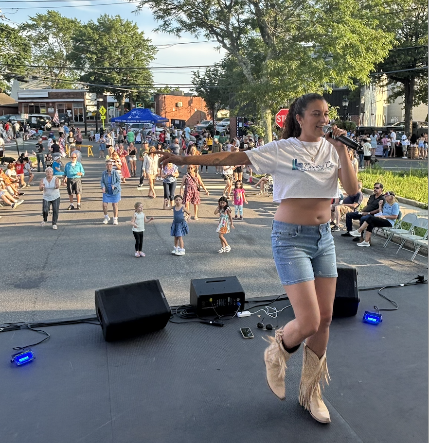 A young woman singing on an outdoor stage with a microphone, wearing a white crop top and denim shorts, under sunny weather, with a crowd of people including children dancing in front of her.
