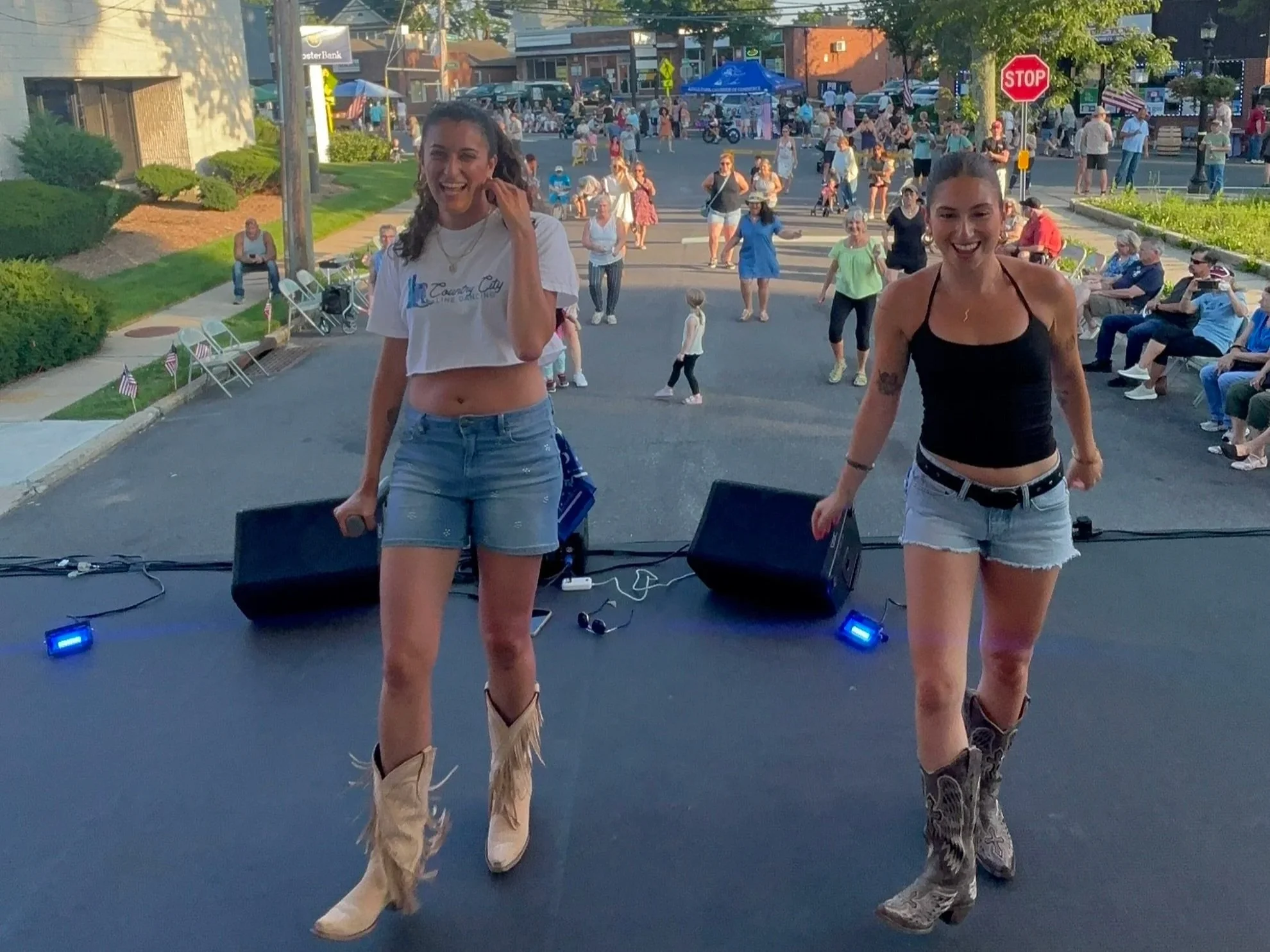 Two women in cowgirl boots and shorts line dance on a stage at an outdoor event and instruct the crowd, with a crowd of people socializing and line dancing in the background.
