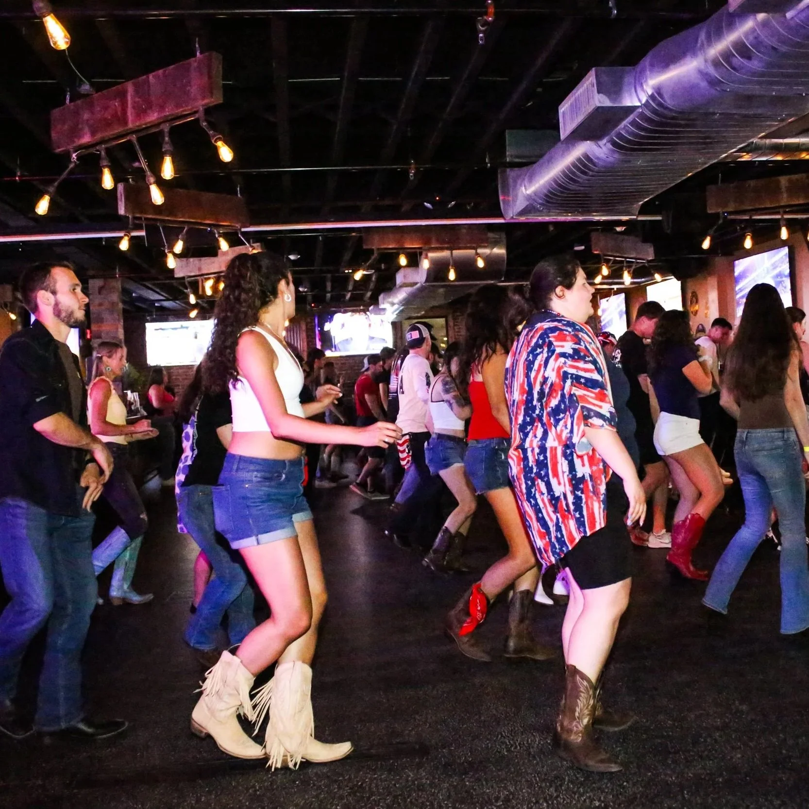 People participating in a line dance or country-style dance inside a dimly lit bar or club, wearing casual and country-themed attire, with wood and brick decor, industrial ceiling pipes, and multiple screens on the walls.